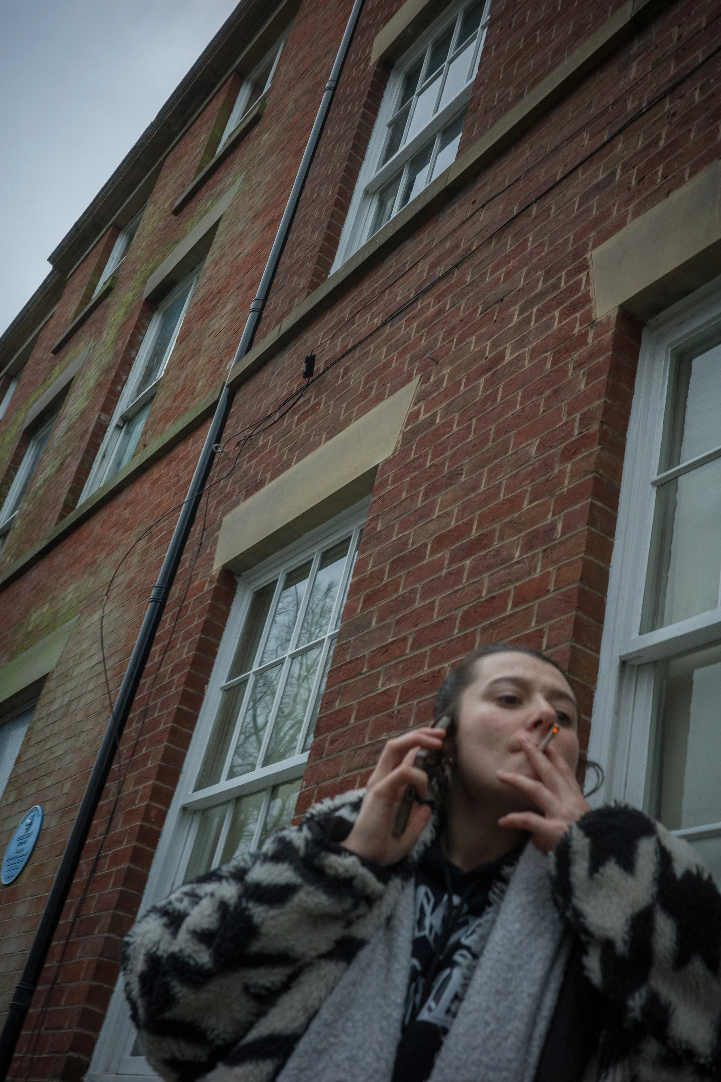 A young woman with long dark hair, wearing a black and white zebra-patterned jacket, is standing outside a brick building. She is holding a phone to her ear and smoking a cigarette, with a focused expression.