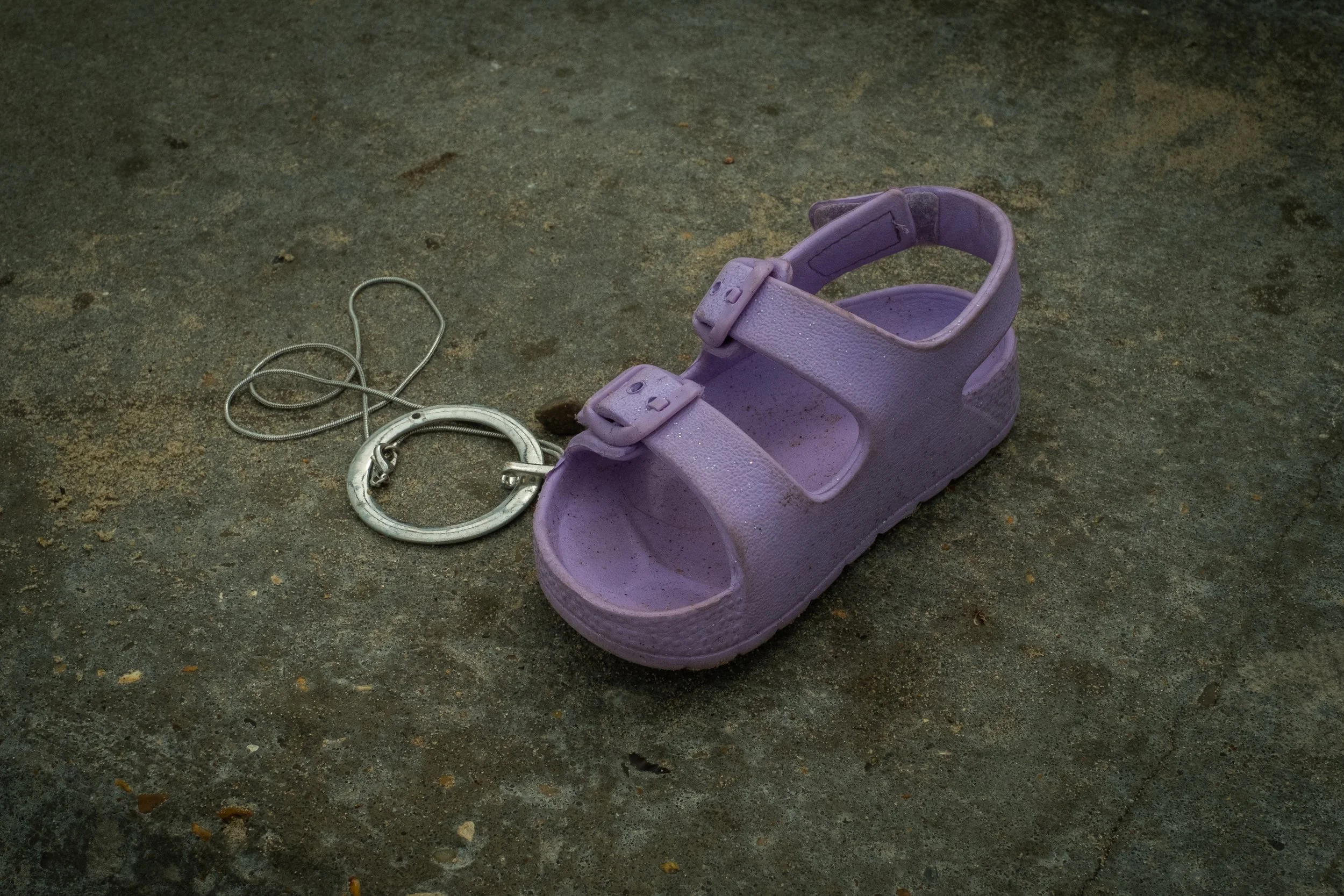 A purple plastic sandal with Velcro straps and a silver hoop earring with a chain on a rough concrete surface.