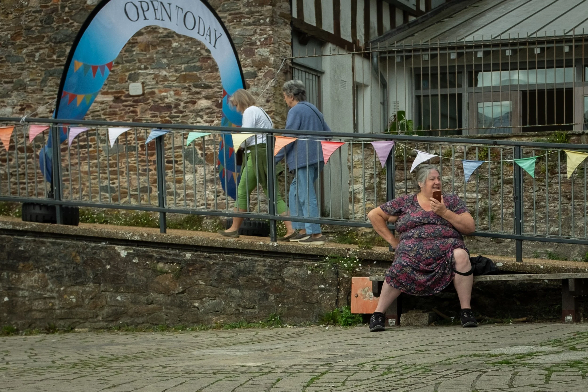 People walking up a small external staircase with a woman sitting at the bottom, all under colorful bunting and a blue arch that says 'Open Today,' against a stone wall.
