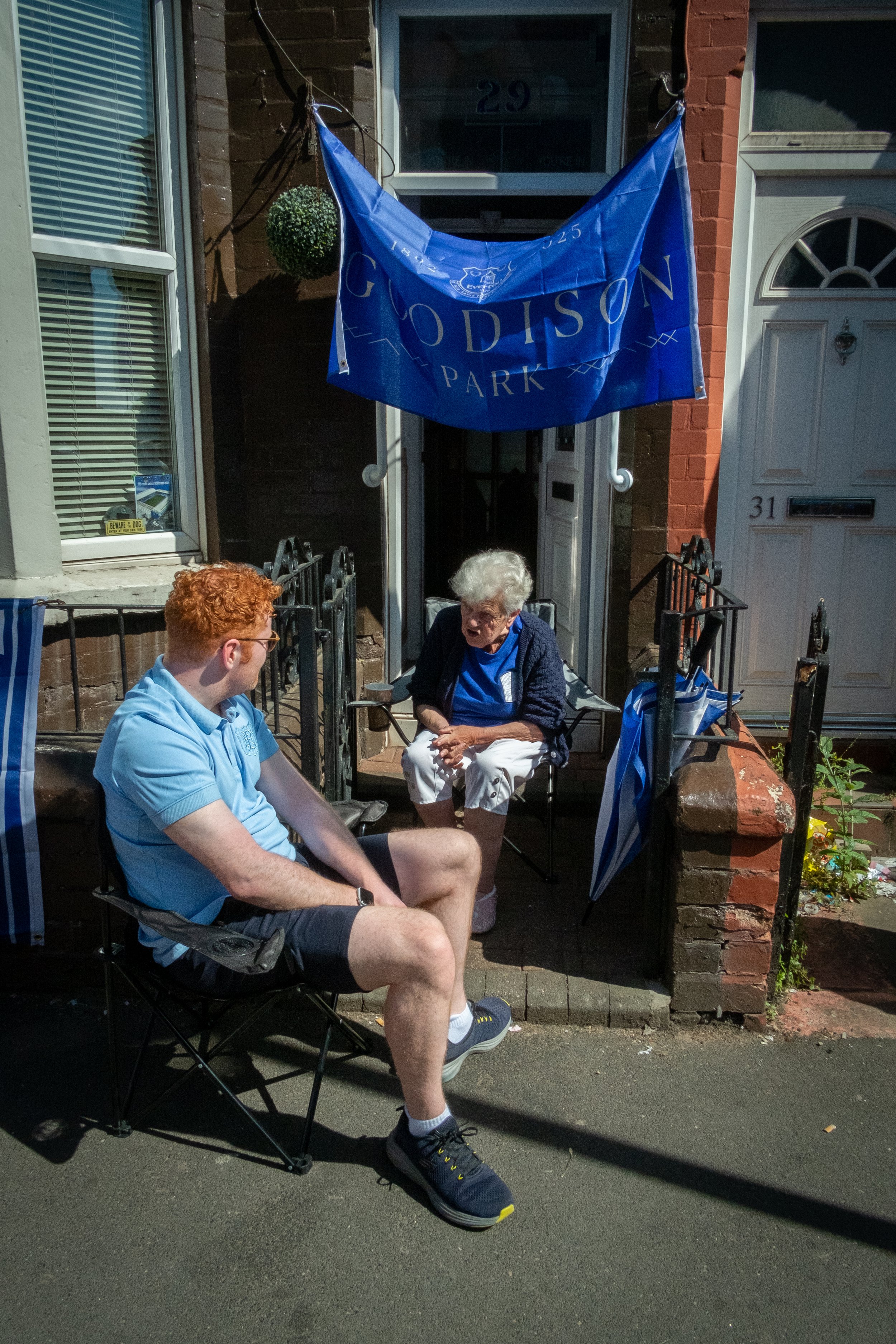 A young man and an elderly woman sit and talk on a front porch decorated with blue and white flags and banners for Goodison Park, a football stadium.