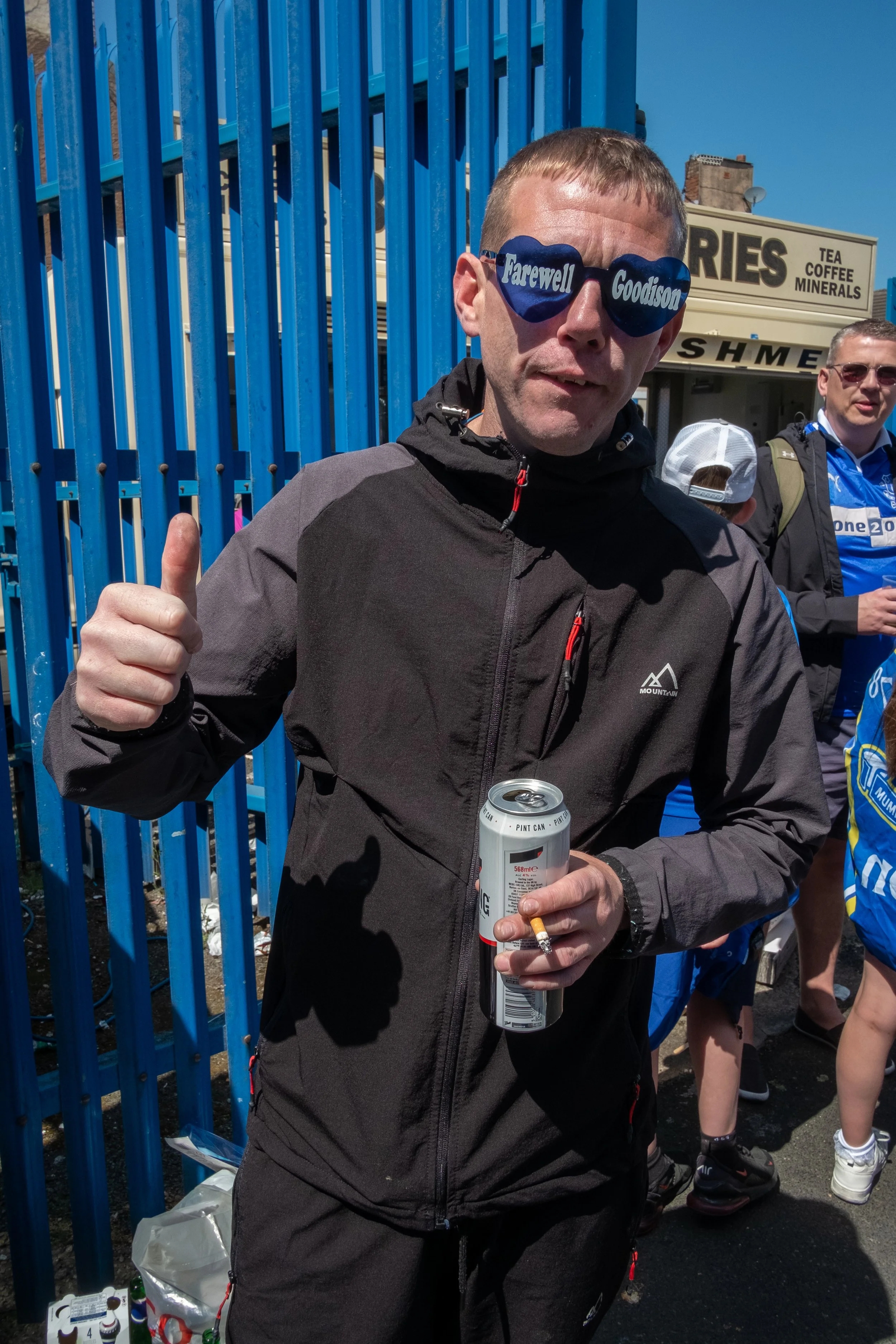 A man holding a beer can and smoking a cigarette, giving a thumbs-up, wearing sunglasses with words 'Farewell' and 'Goodison' on the lenses, and dressed in outdoor gear, at an outdoor event with a blue fence and a food stand in the background.