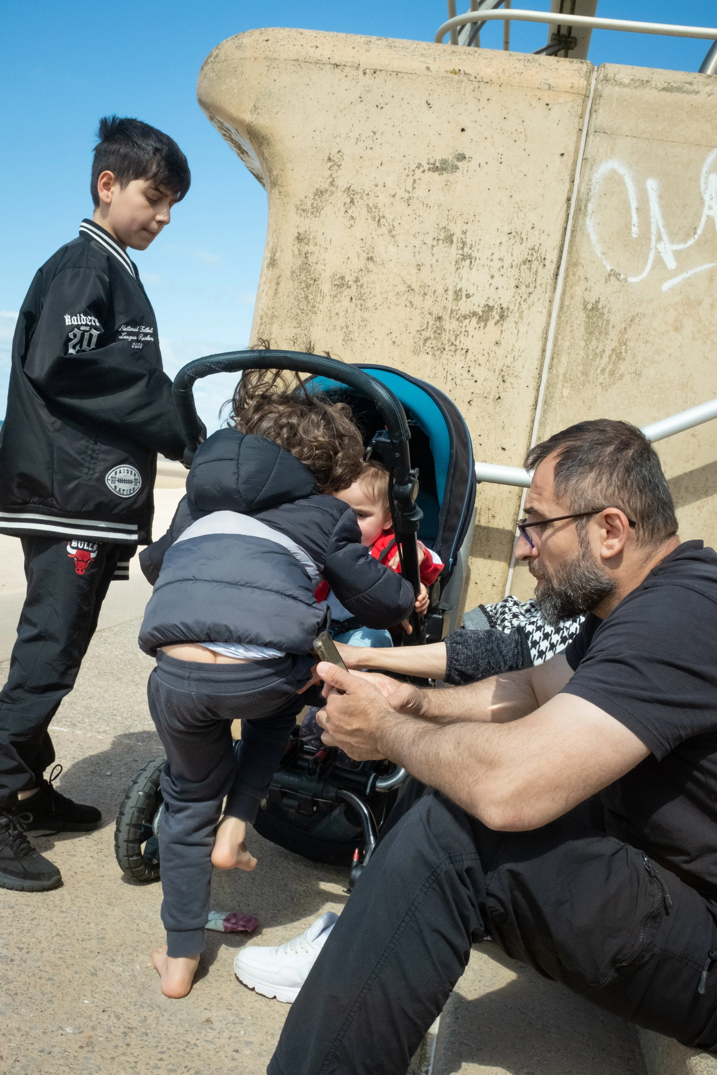 A young boy standing and a man sitting on the ground, both interacting with two children near a stroller on a sunny day, with a concrete structure and blue sky in the background.