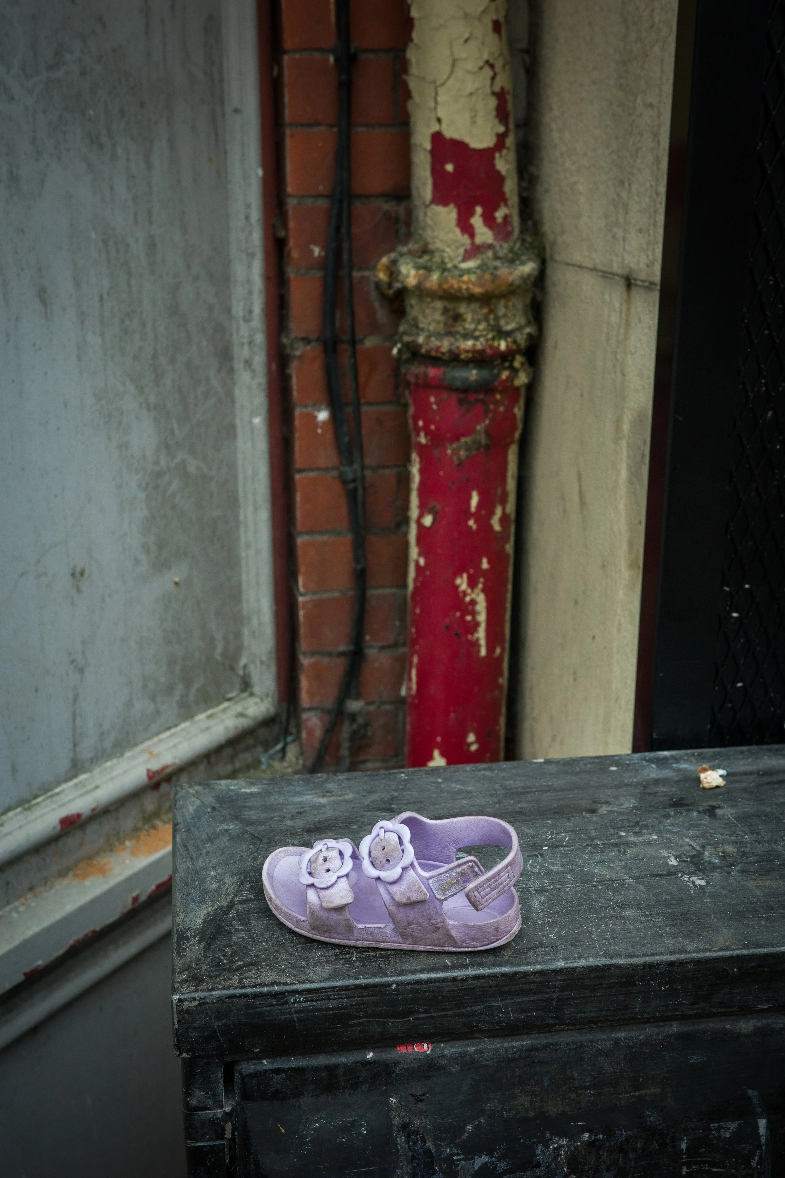 A pair of small purple children's sandals with bear and flower decorations on a black, weathered table near a brick and concrete wall with a red, rusted pipe.
