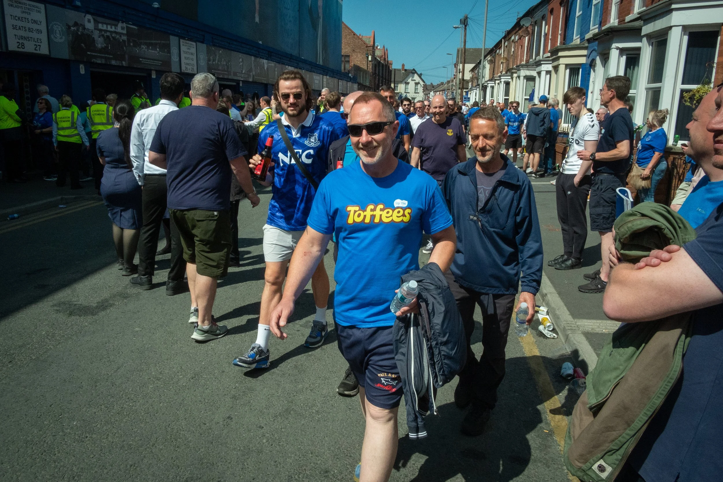 Crowd of sports fans walking on a city street during a daytime event, some wearing Everton football club jerseys and holding drinks.