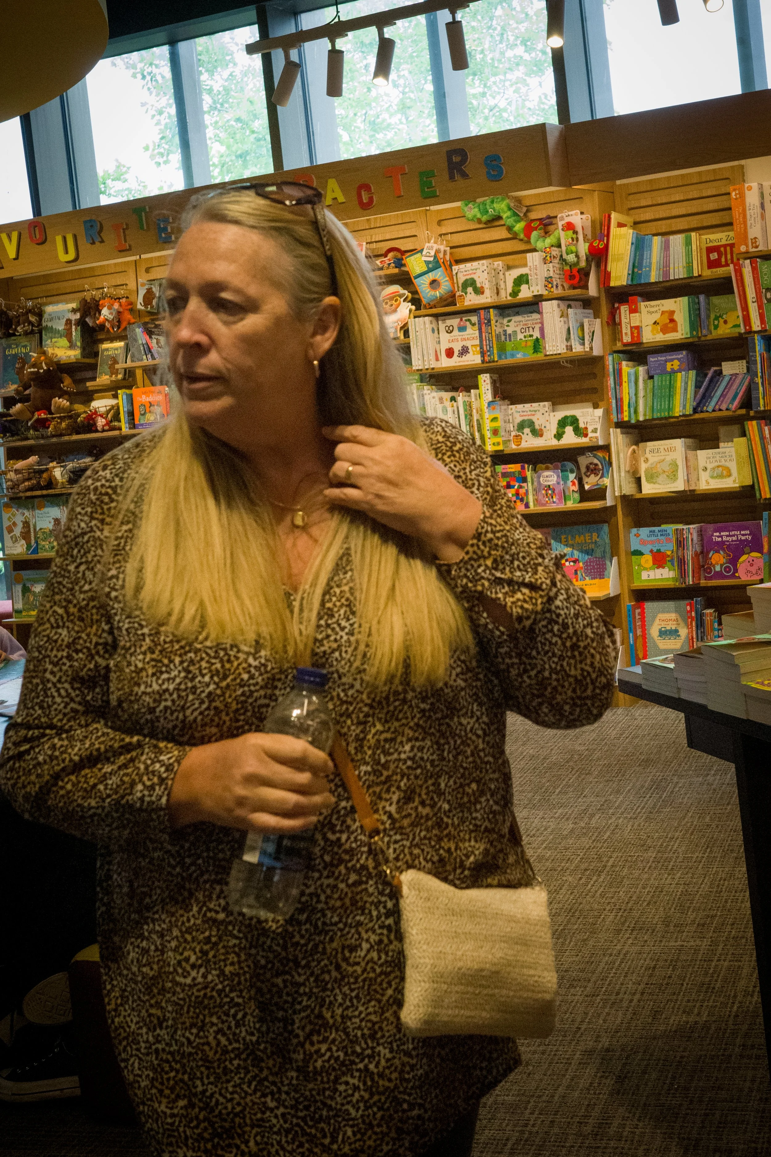 A woman with long blonde hair wearing a leopard-print top and sunglasses on her head, holding a water bottle, standing in a bookstore with children's books on the shelves behind her.