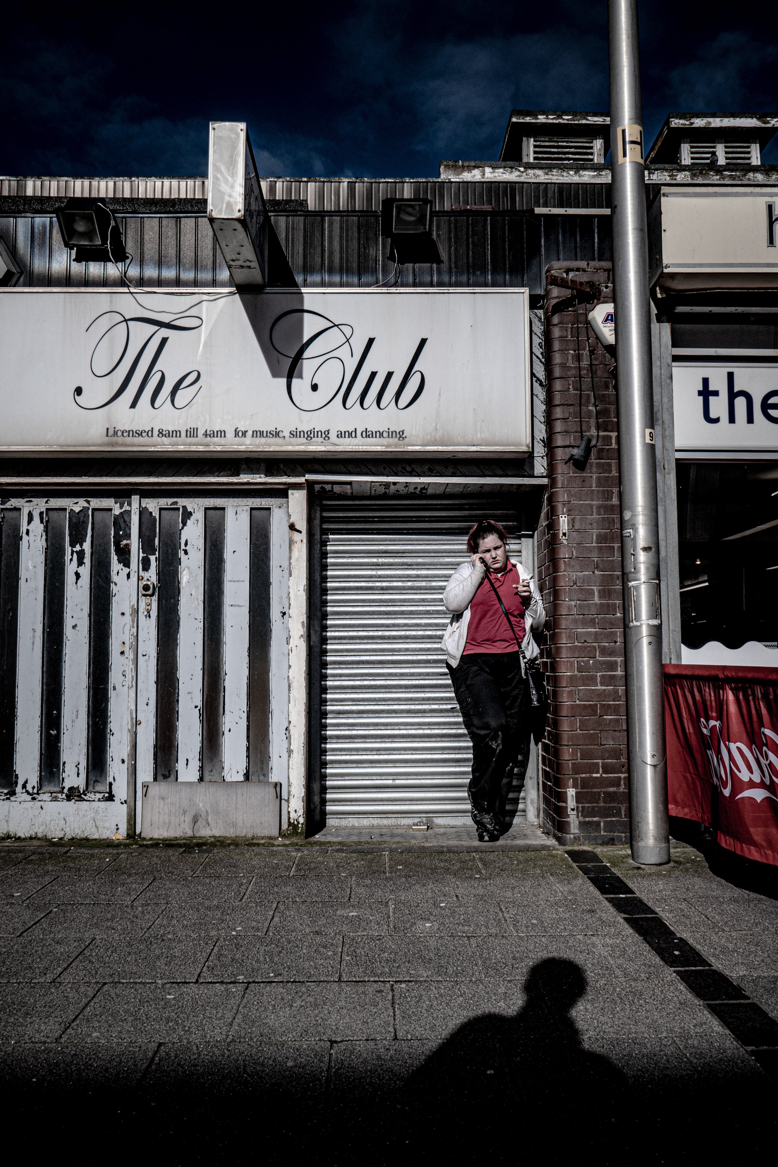 A woman with short hair in a red jacket and black pants stands outside a closed shop with a metal roller shutter, talking on her cellphone. There is a sign above the shop entrance that reads 'The Club' with hours of operation from 8am to 4am, and a p