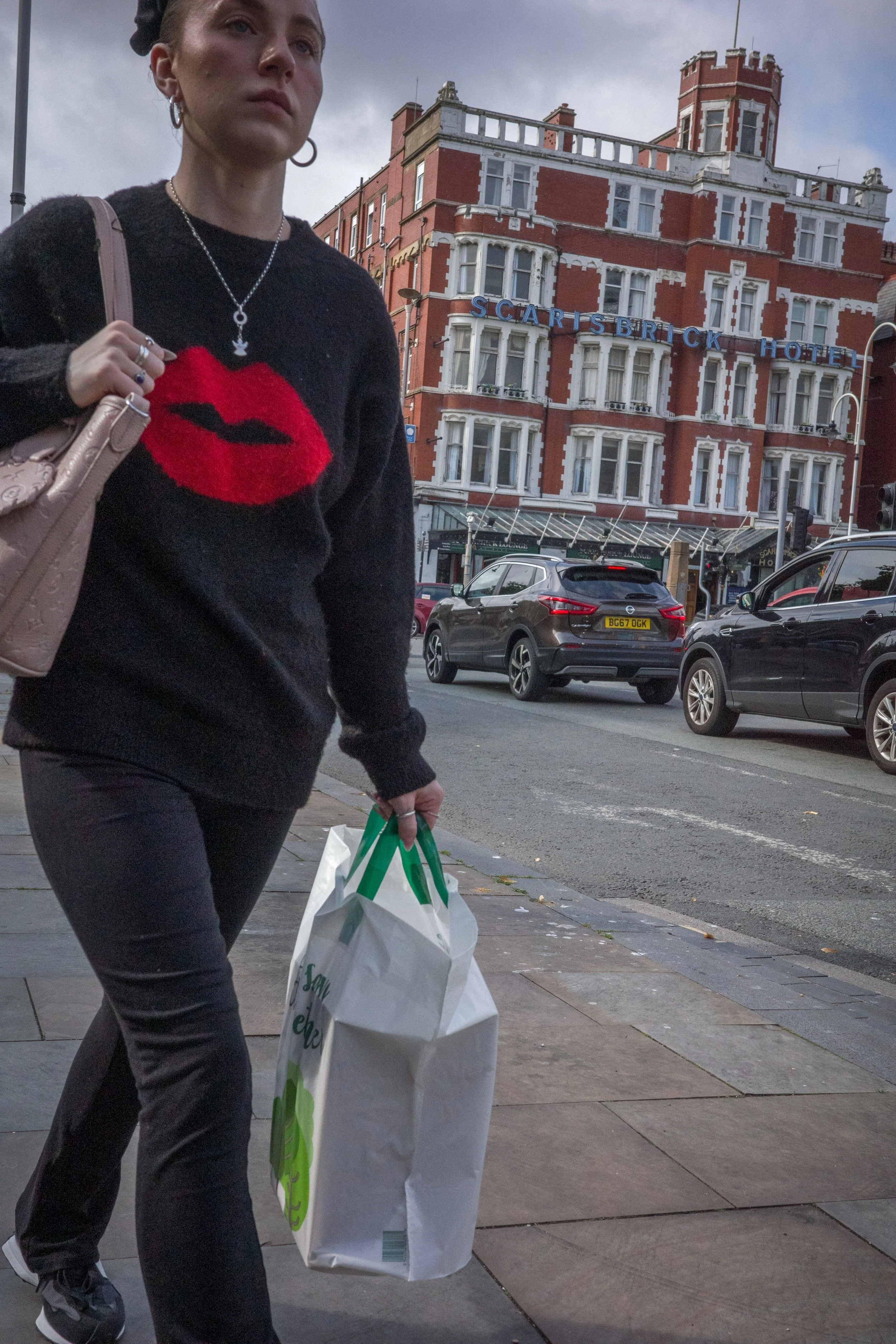 A woman walking on a city sidewalk carrying a shopping bag, in front of a red-brick hotel building with a sign that reads "Scarisbrick Hotel."