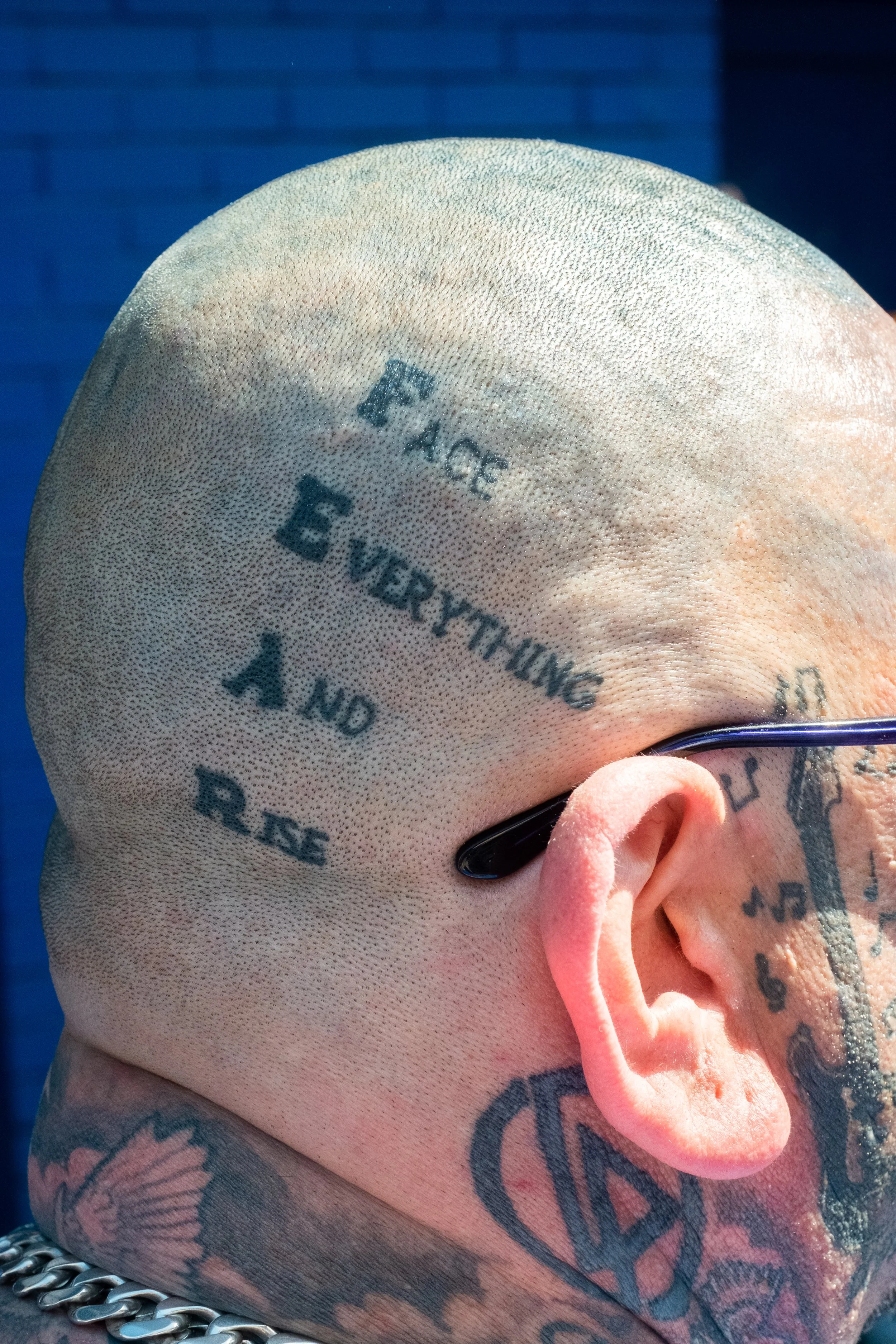 Close-up of a person's shaved head with tattoos, including a quote and symbols, wearing sunglasses and a chain necklace, against a blue background.
