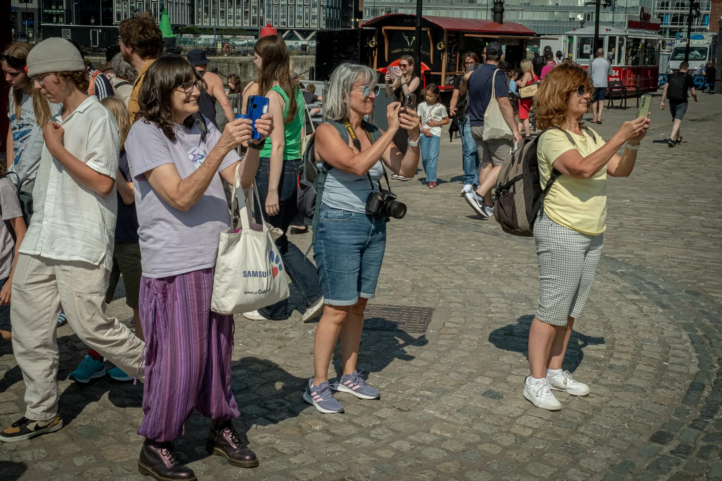 A group of people standing on a cobblestone waterfront, taking photos and selfies, with boats and modern buildings in the background.