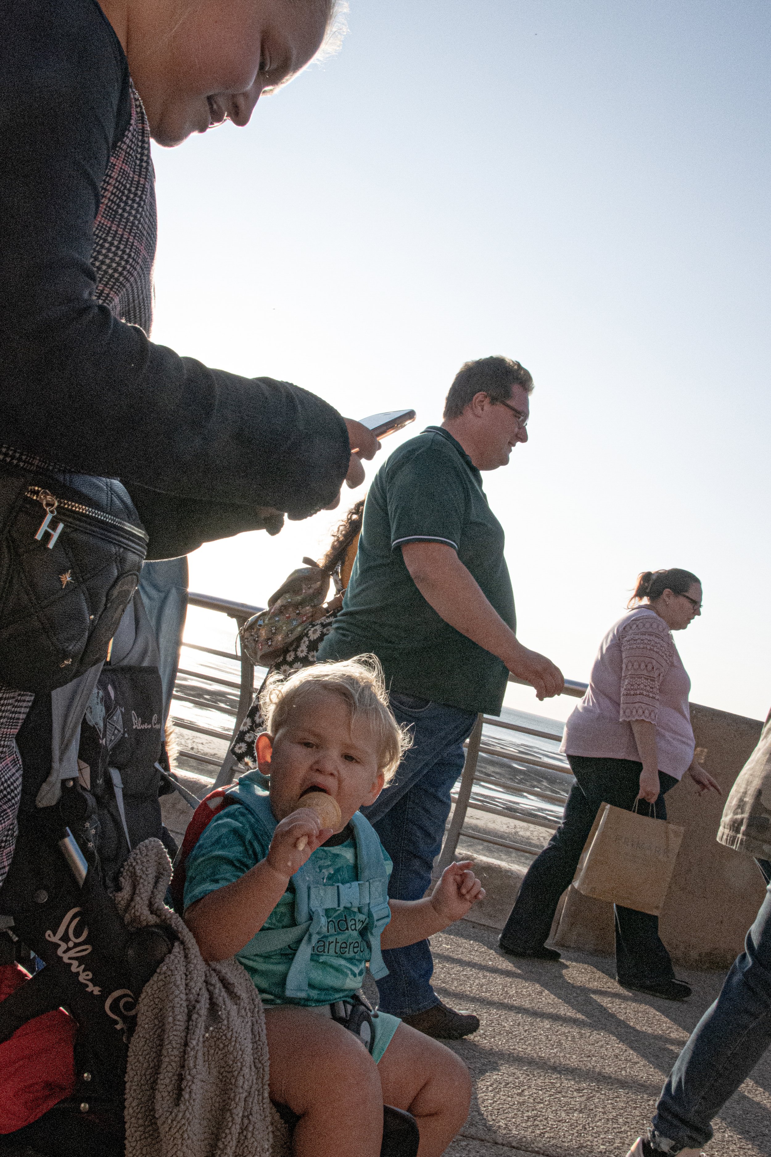 A young boy with curly red hair, seated, wearing a turquoise shirt and a harness, eating an ice cream cone, with a tan towel and red backpack nearby, at a place with people walking on a boardwalk against a backdrop of a beach and ocean at sunset.