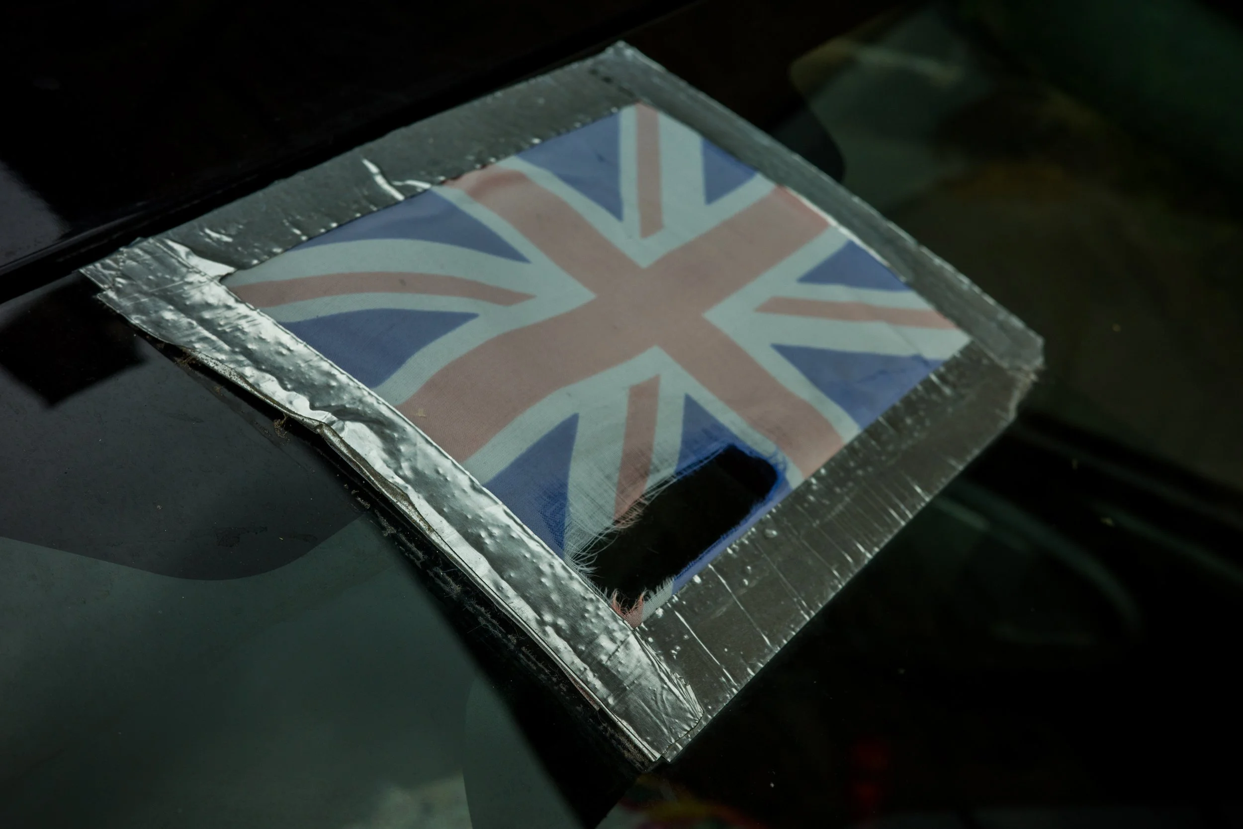 Close-up of a printed Union Jack flag under a clear plastic sheet, secured with metallic tape, on a black surface.