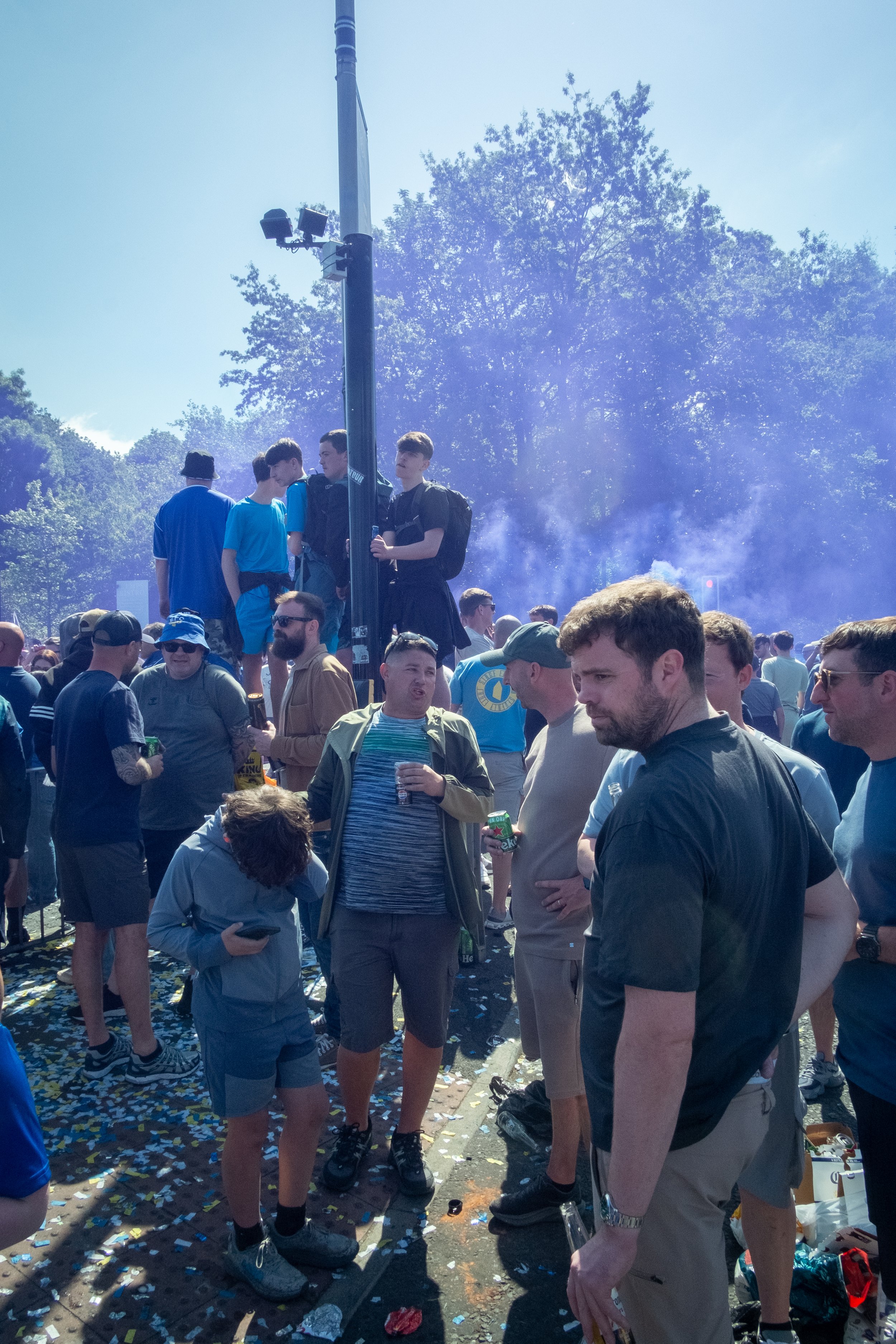 Crowd gathered outdoors during a celebration or festival, with some people standing on a platform and others on the ground, amidst confetti and smoke, and surrounded by trees and blue sky.