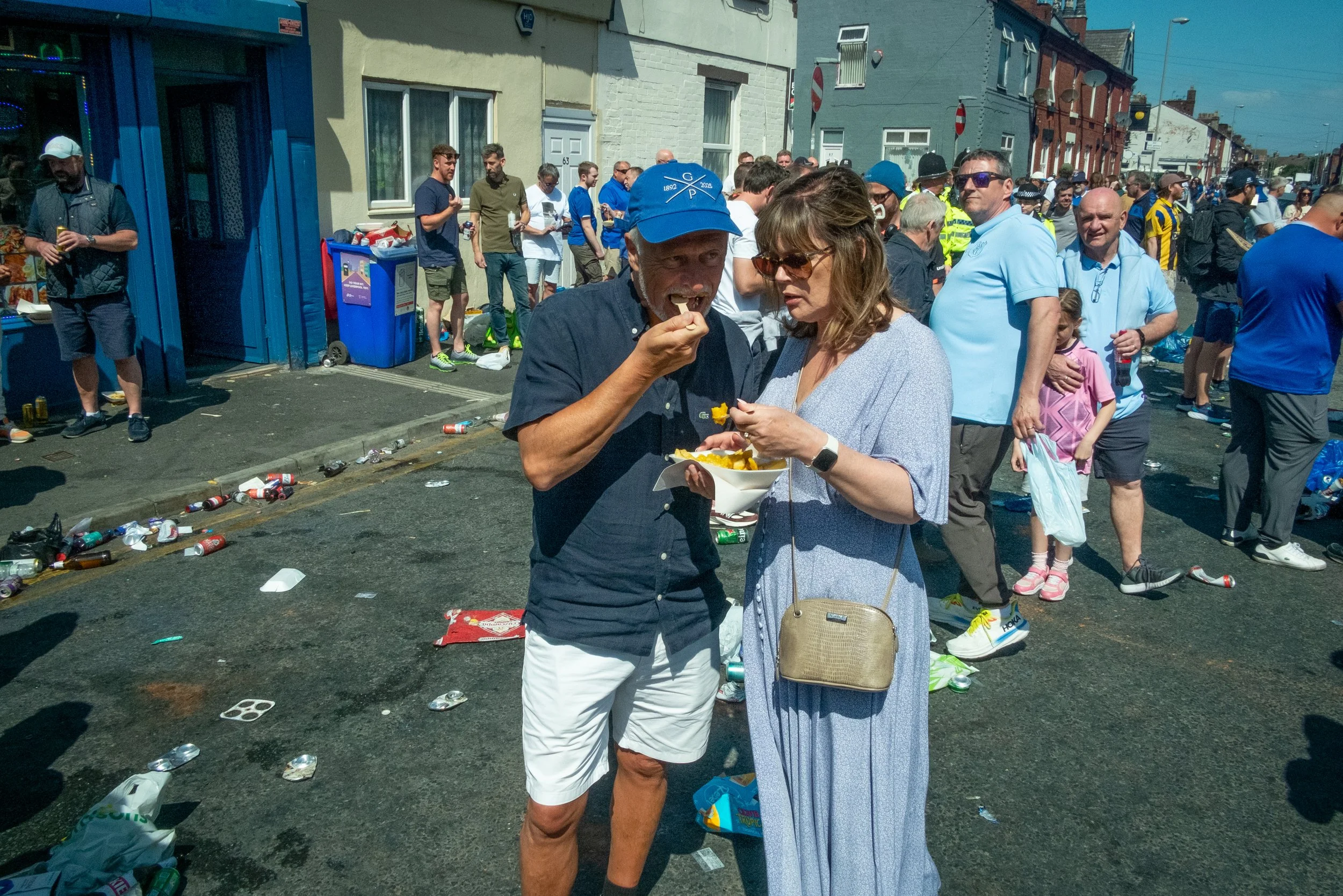 Two people eating chips at a crowded street party with littered street and groups of people in the background.