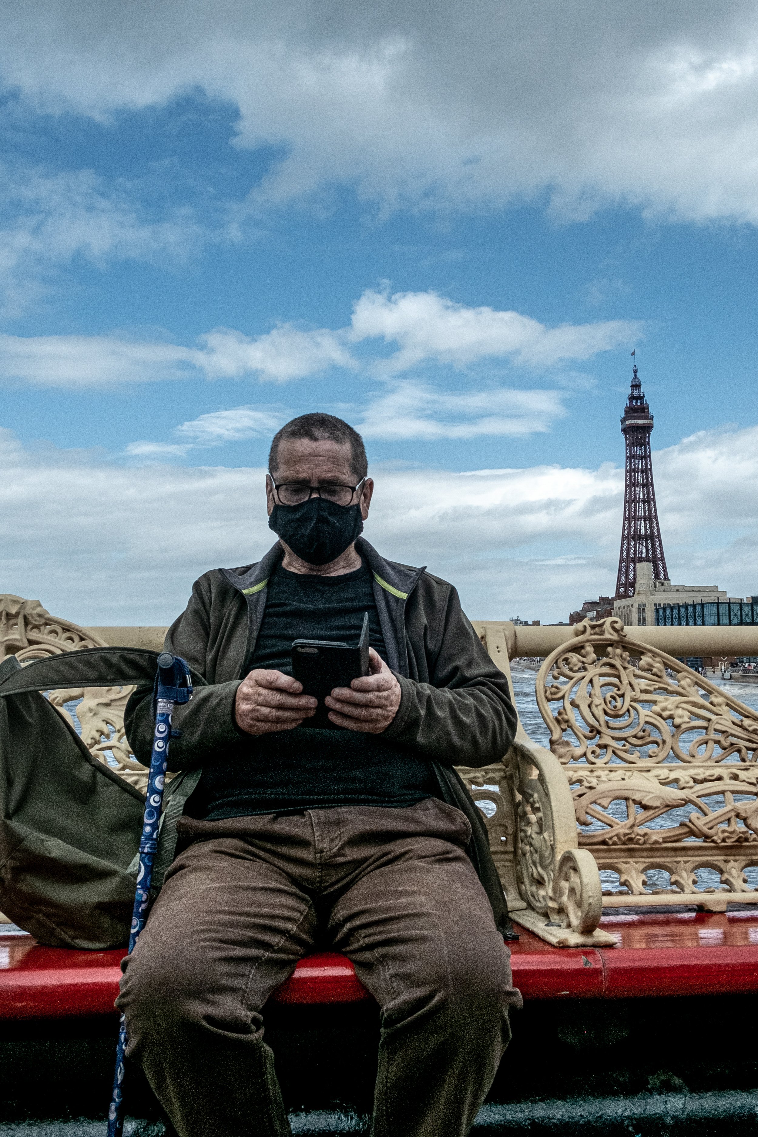 A man wearing glasses and a black face mask, sitting on a decorative bench with the Tokyo Tower in the background.