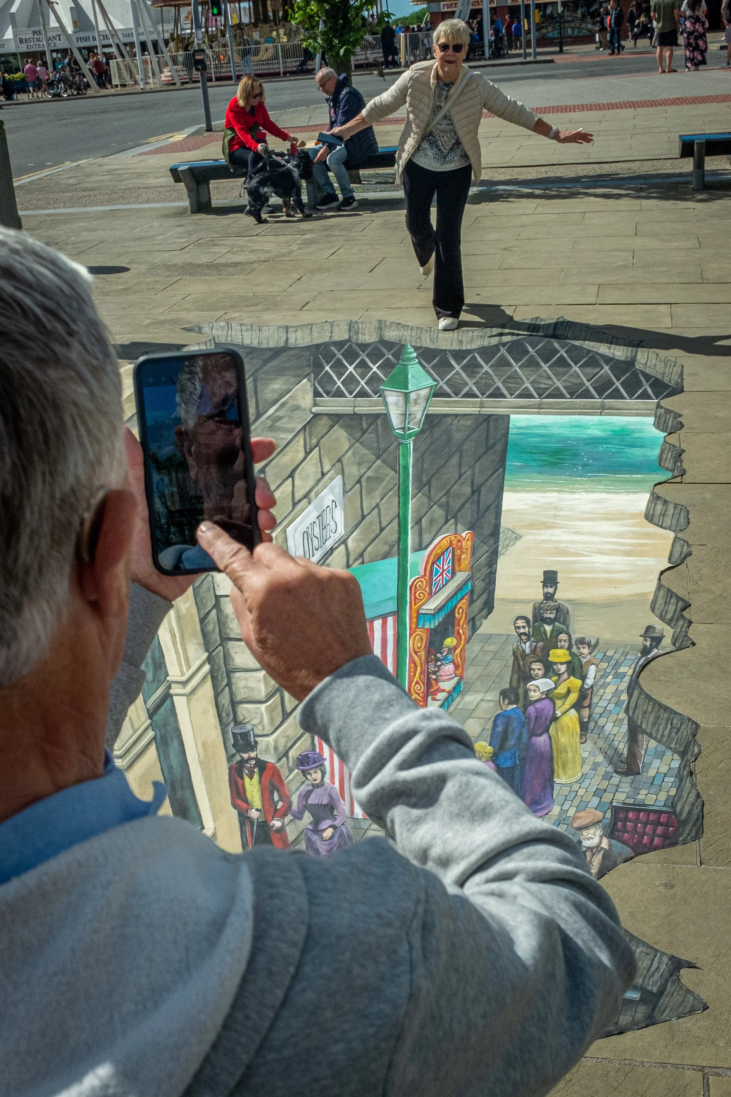 A street performer uses a painted cutout of a city scene to create a 3D illusion of a hole in the sidewalk, showing a scene with people, a lamp post, and buildings. An elderly woman in a beige jacket and sunglasses appears to be stepping out of the h
