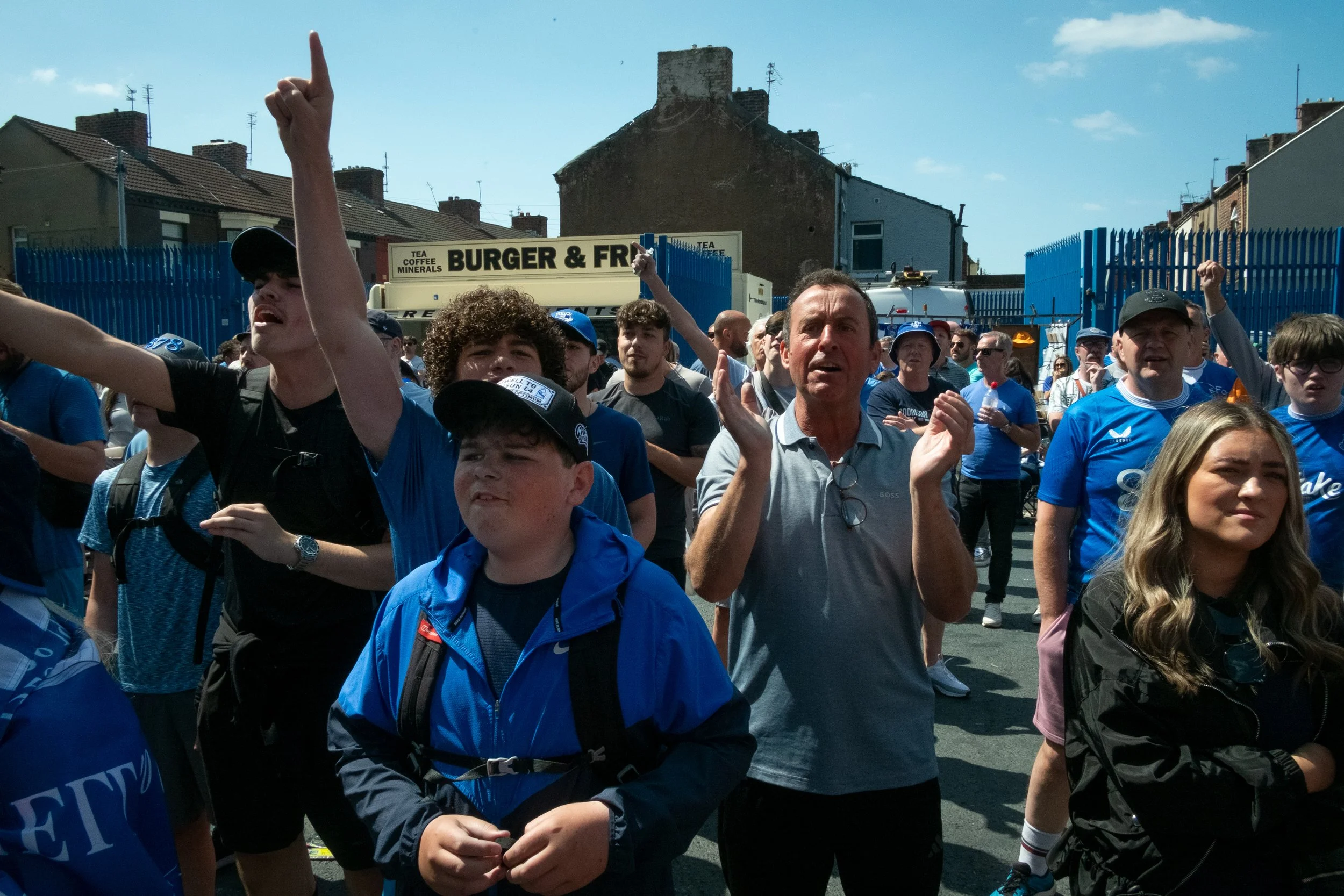 Group of people, including children and adults, gathered outdoors, with some raising their fists or clapping, in front of a blue fence and a sign reading "Burger & F**" on a sunny day.