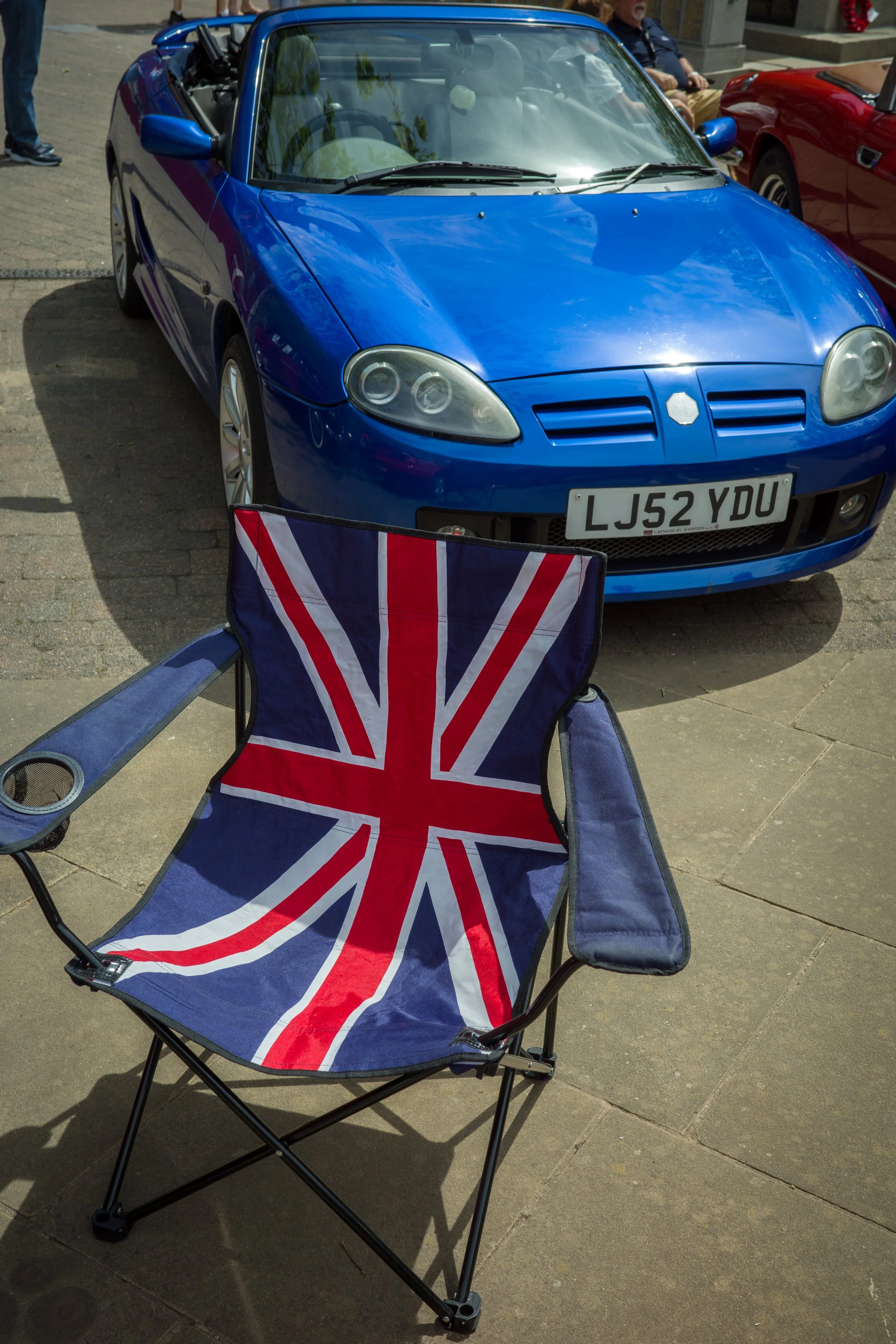 A blue convertible sports car parked on a street, with a Union Jack flag patterned folding chair in the foreground.