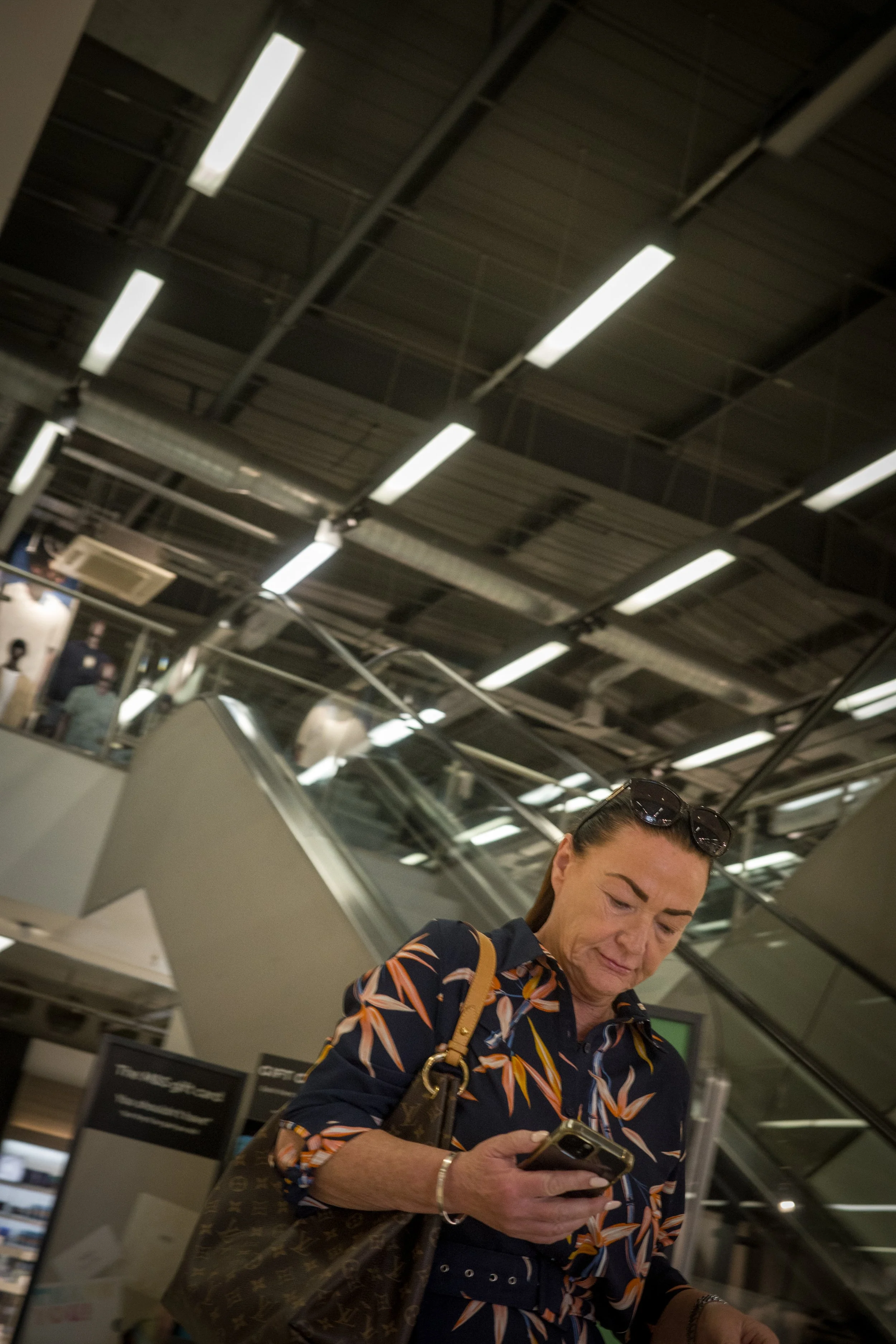 A woman in sunglasses and a patterned shirt looking at her phone in an indoor setting with overhead lights, escalator, and a newsstand.