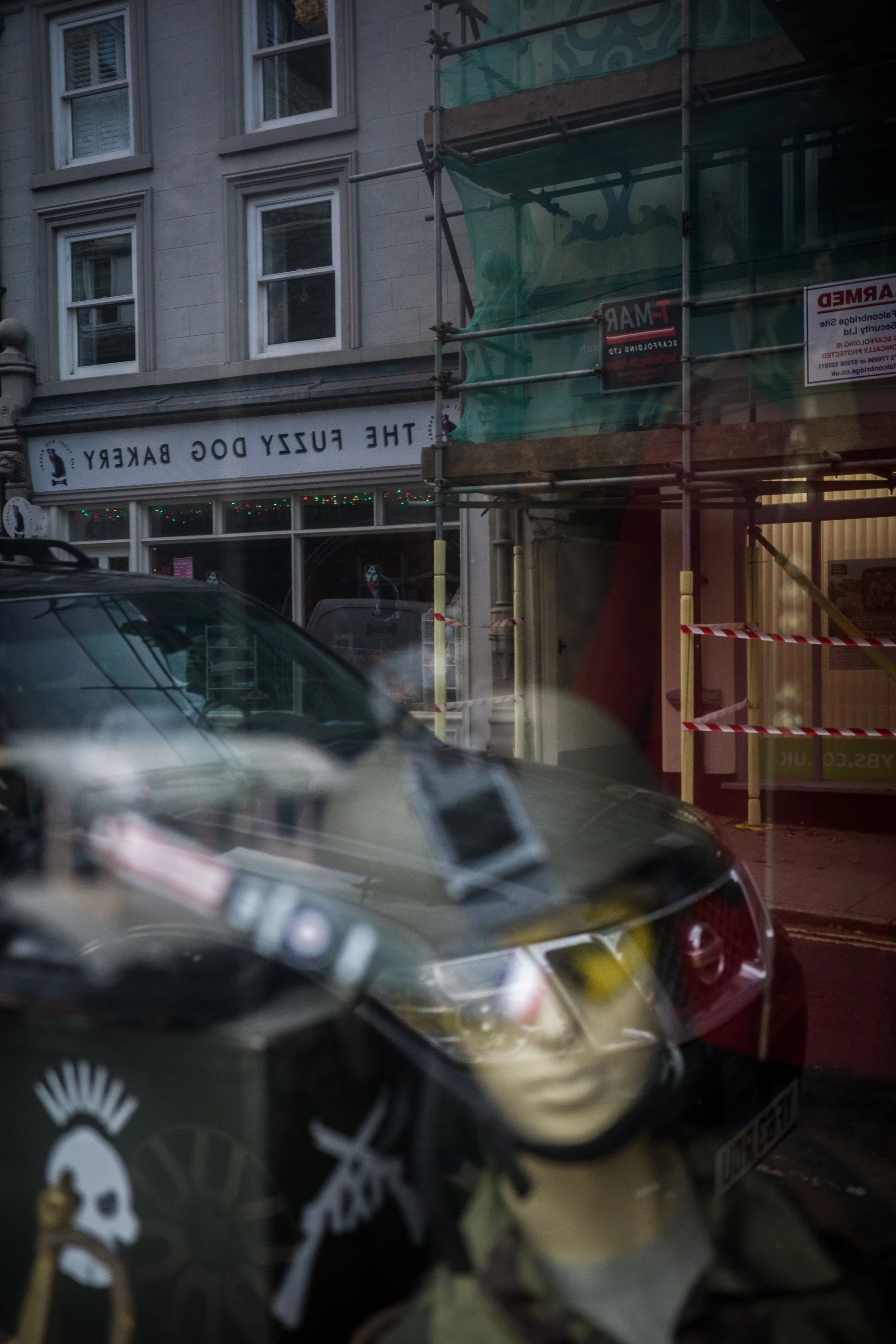 Reflection in a glass window showing a street scene with a car, a building with windows, and a storefront named "The Fuzzy Dog Bakery". There is also scaffolding outside the building.