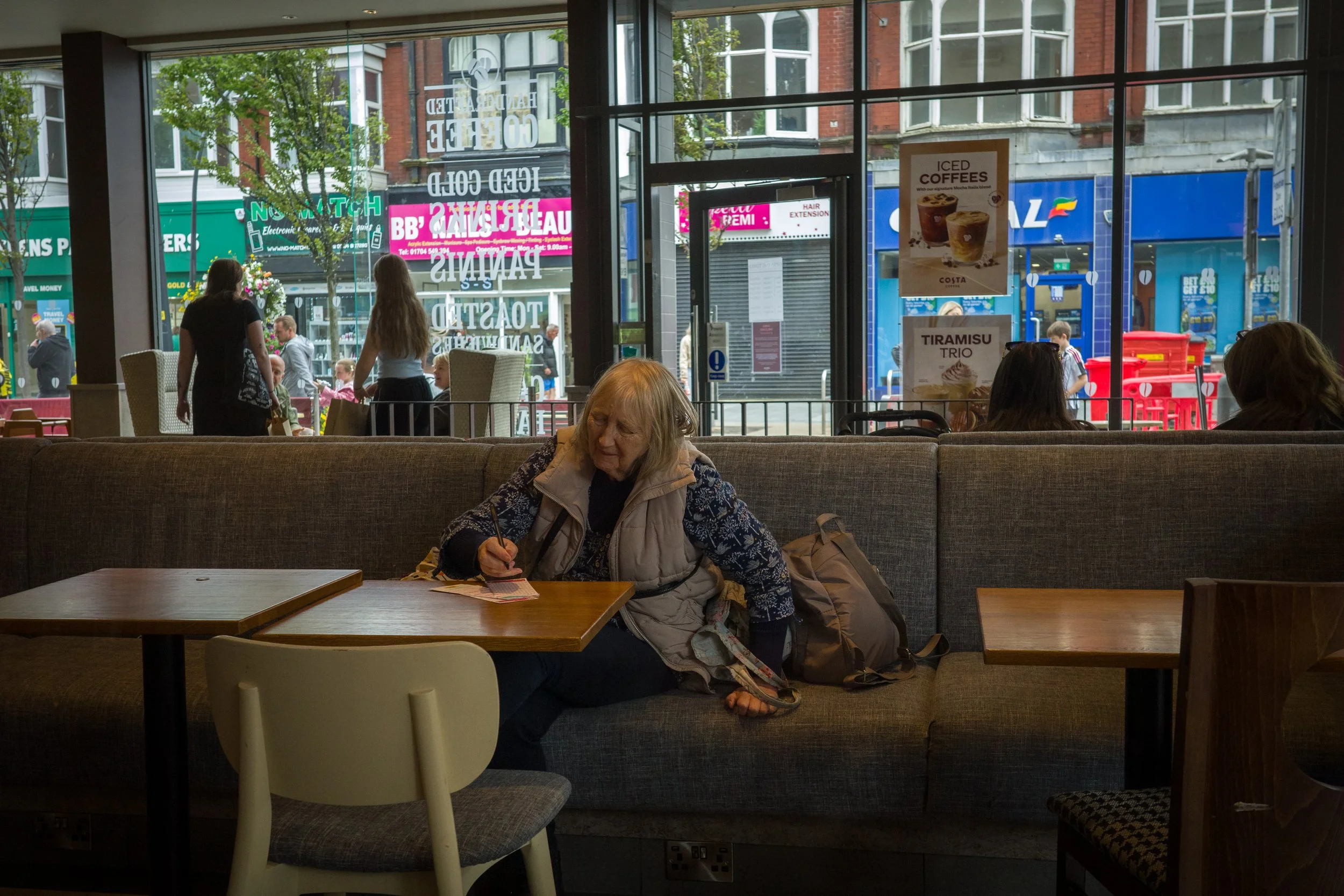An elderly woman sitting on a gray cushioned bench inside a cafe, writing on a small card. Outside the cafe's large glass windows, pedestrians walk along a busy storefront-lined street with signs and advertisements.