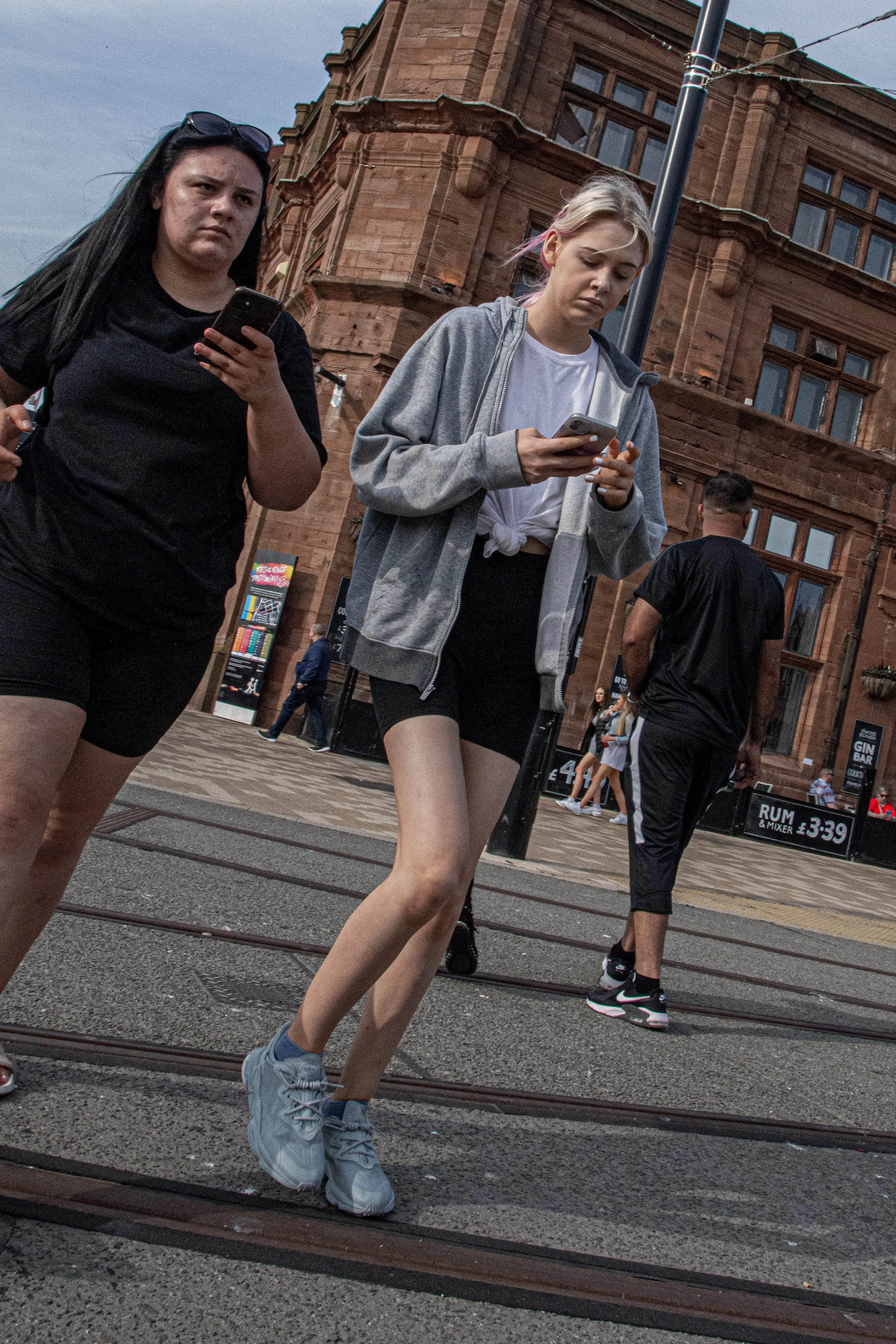 Two young women walking on a street, focused on their smartphones. One has long black hair, sunglasses on her head, and is wearing a black t-shirt and shorts. The other has light hair with pink streaks, wearing a grey hoodie over a white t-shirt and 