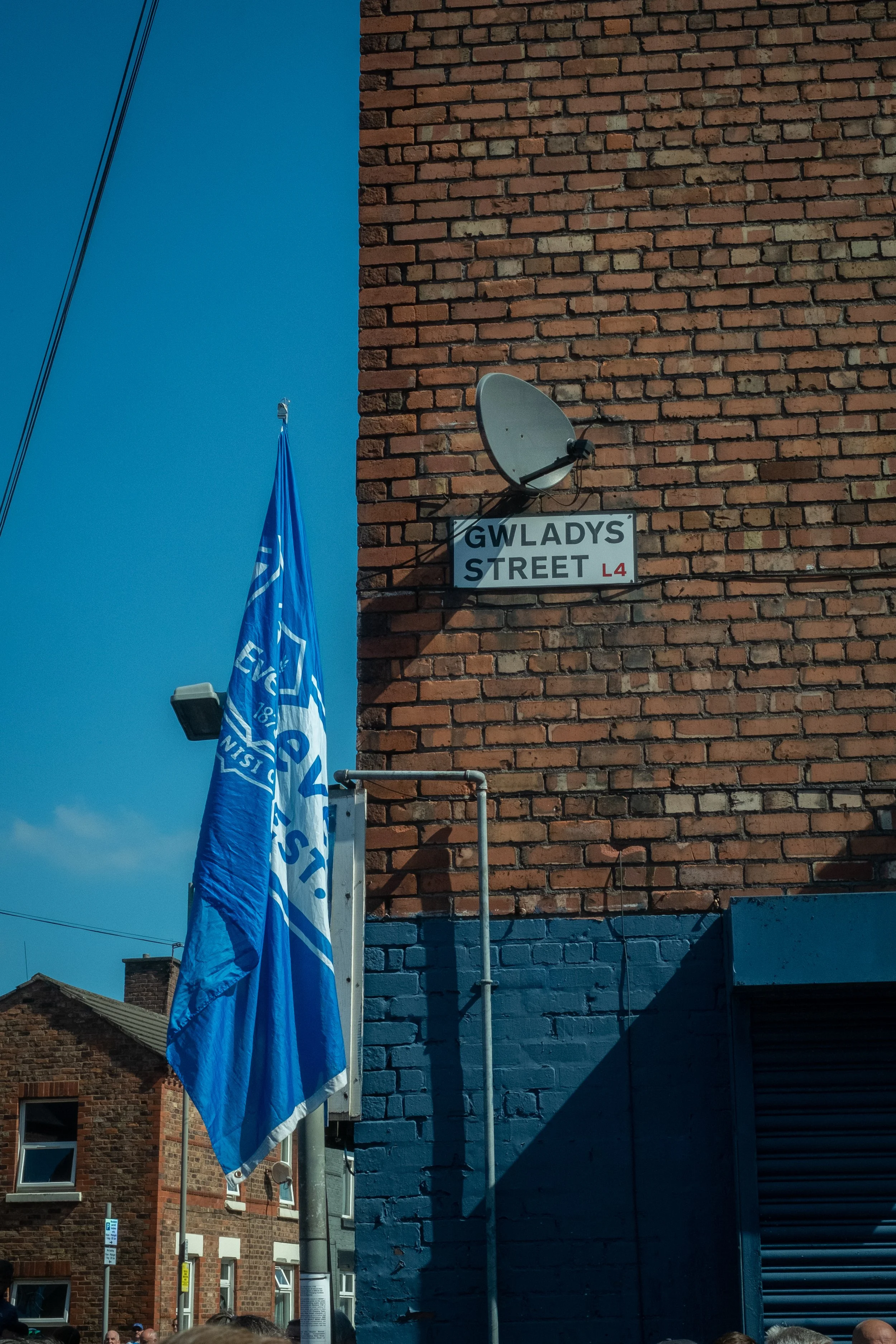 Street corner with a brick building, street sign reading "GWLADYS STREET L4," a satellite dish, a blue flag, and blue painted lower wall, with neighboring buildings and a clear blue sky.