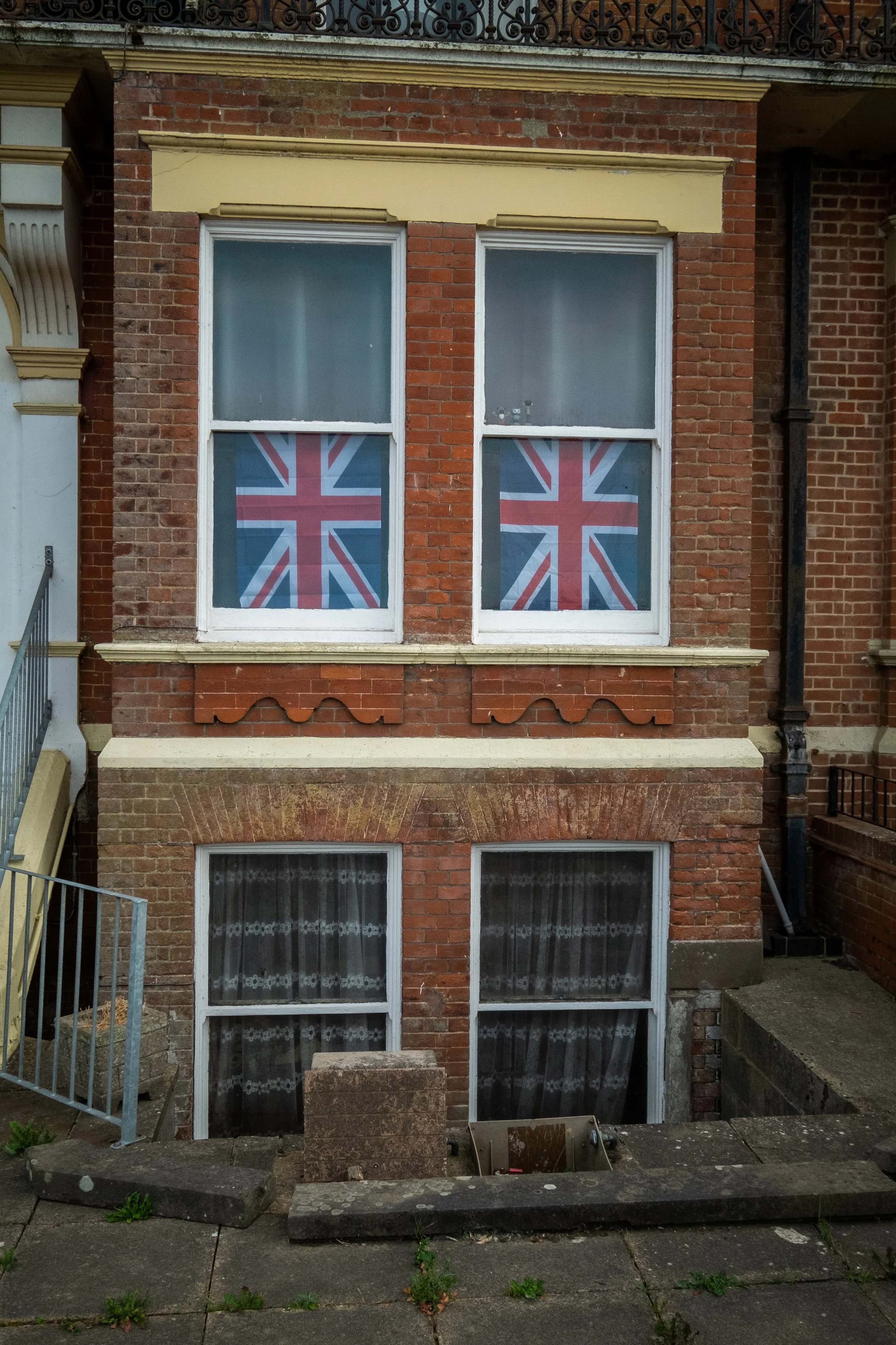 A brick building with two large windows, each decorated with a union jack flag, and a small set of stairs leading down to an entrance. The ground in front of the building has some scattered weeds and sidewalk tiles.