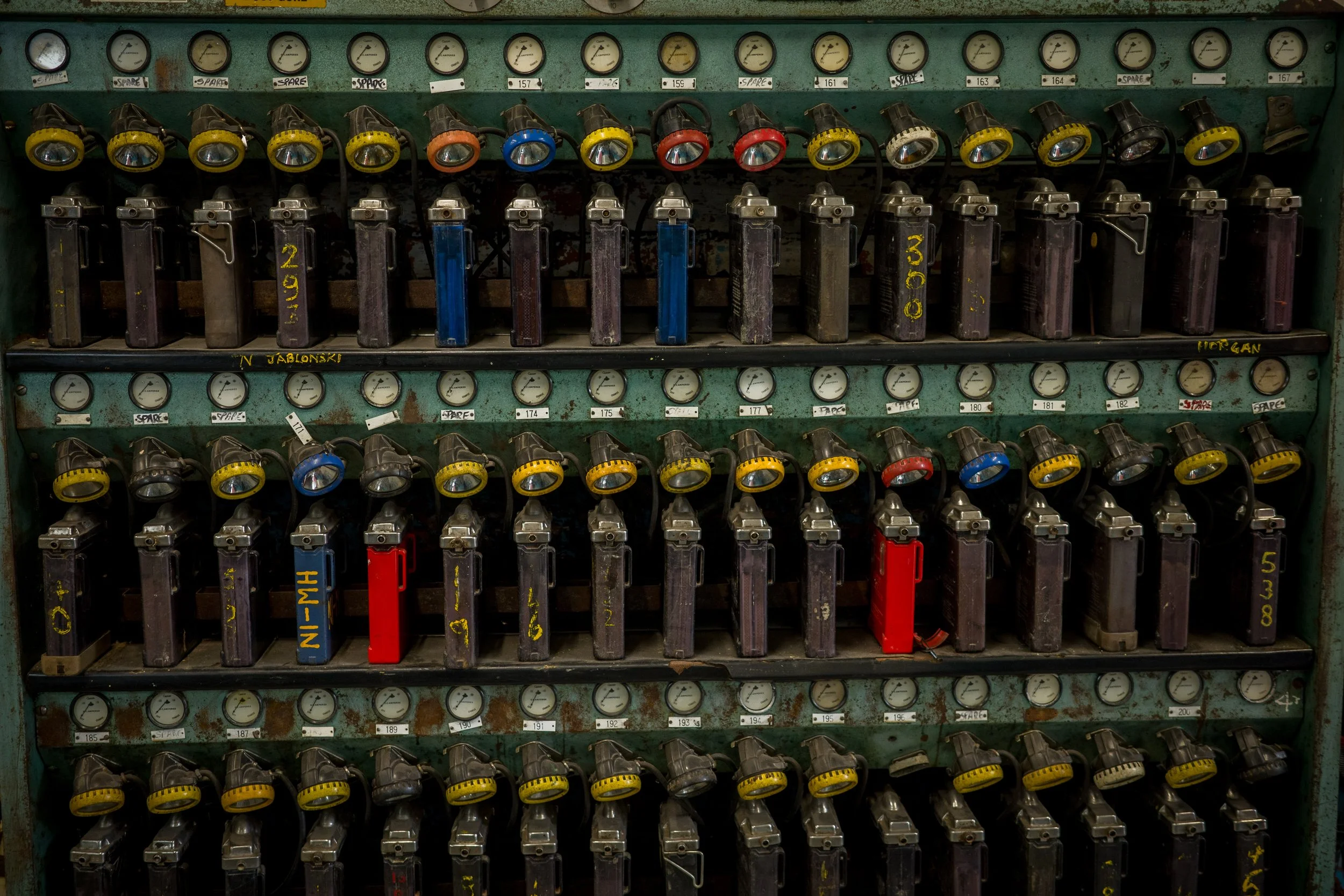 An old green metal control panel with multiple rows of gauges, switches, and colored knobs, some labeled with handwritten tags.