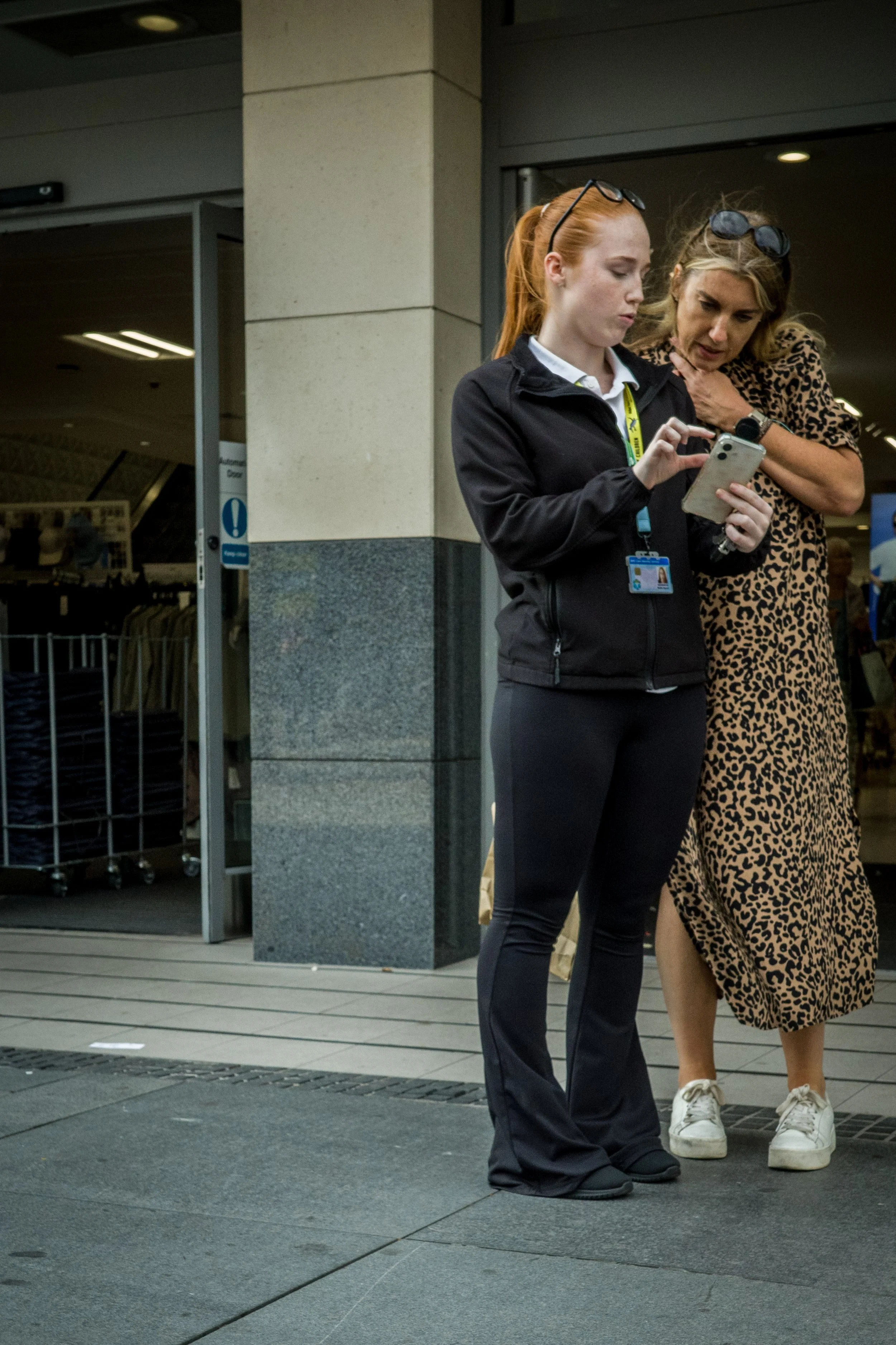 Two women standing outside near an entrance, one is looking at a phone and the other is touching her chest, both wearing glasses on their heads.
