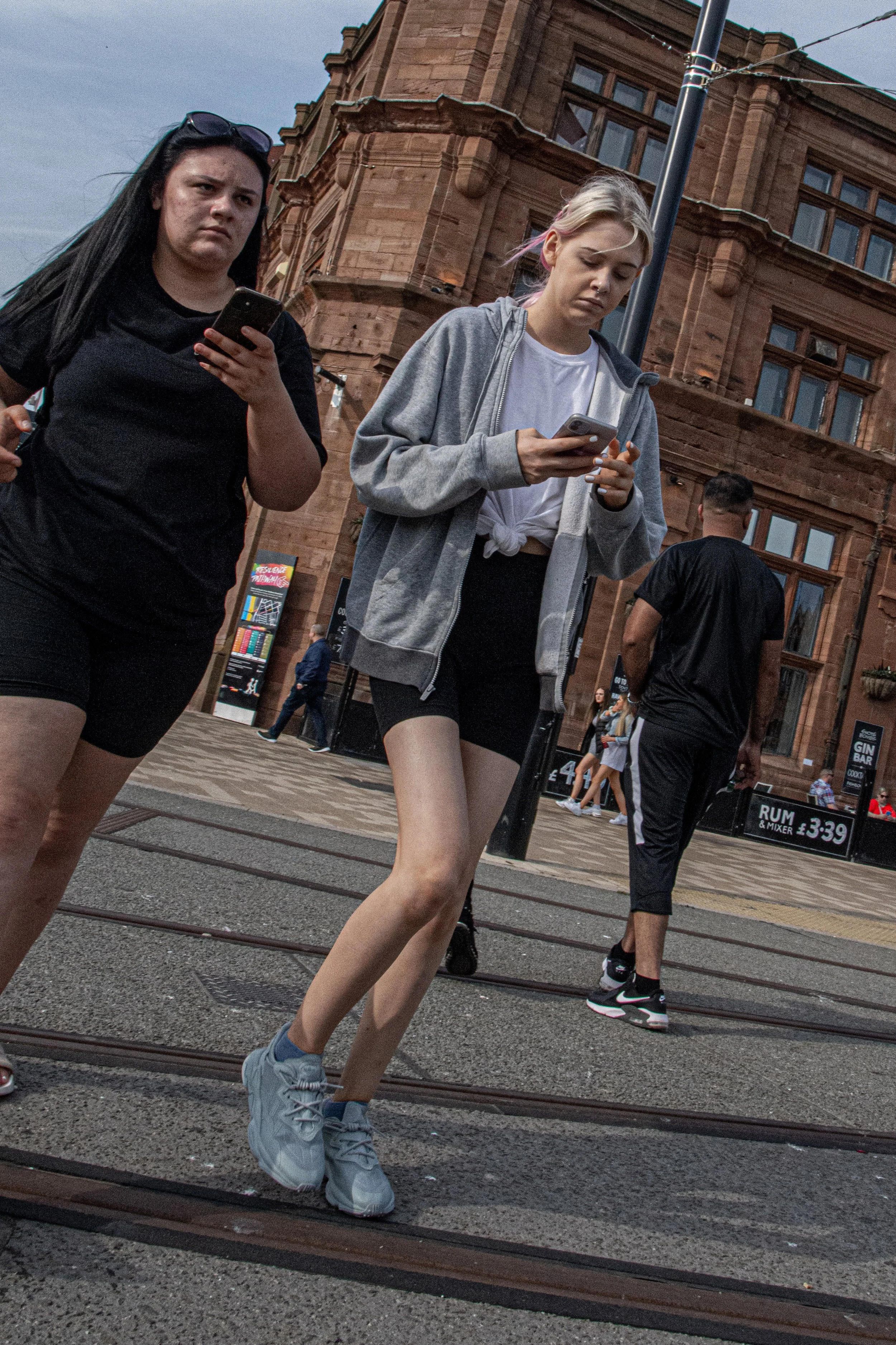 Two young women walking and looking at their phones on a city street with historic red brick building in the background. One woman has long black hair, sunglasses on her head, and is wearing a black t-shirt and shorts. The other has blonde hair tied 