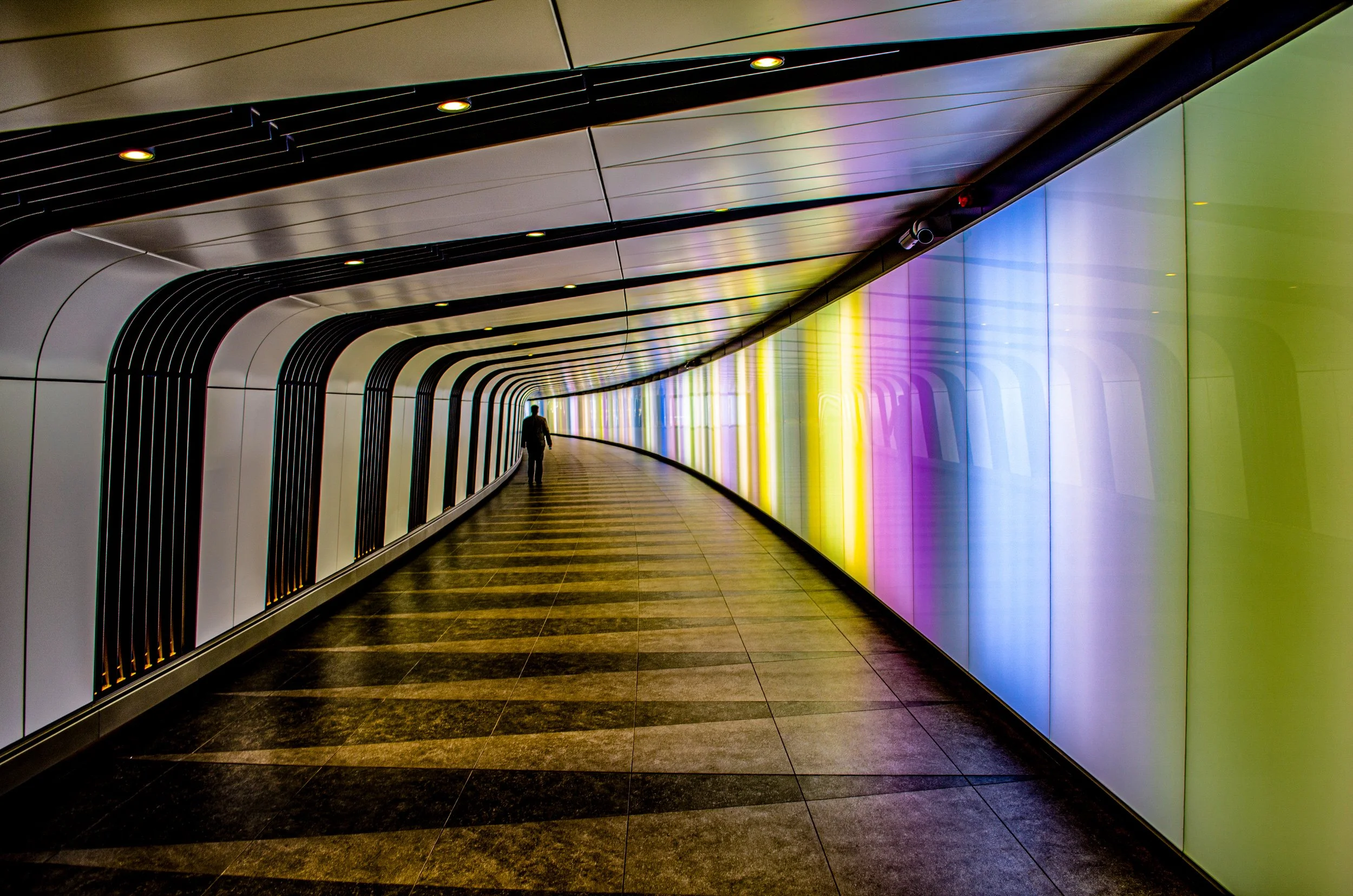 A person walking through a modern, curved corridor with colorful illuminated panels on one side and black-grilled railings on the other.