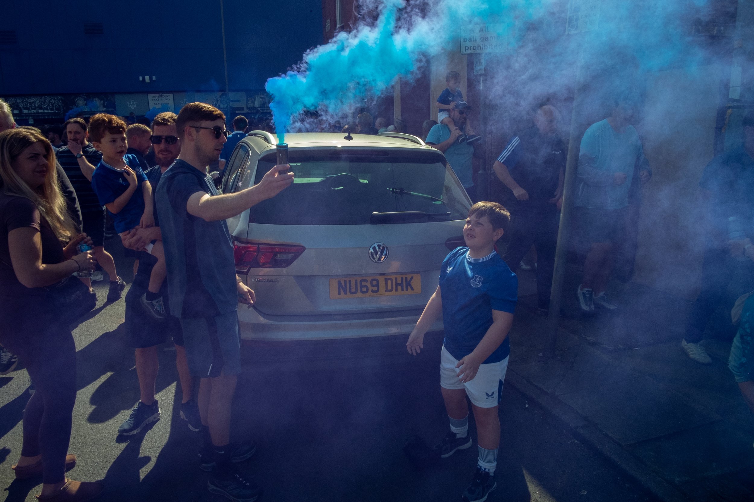 A crowd of people, including children and adults, gathered around a man holding a blue smoke flare in front of a white Volkswagen car, with blue smoke billowing into the air.