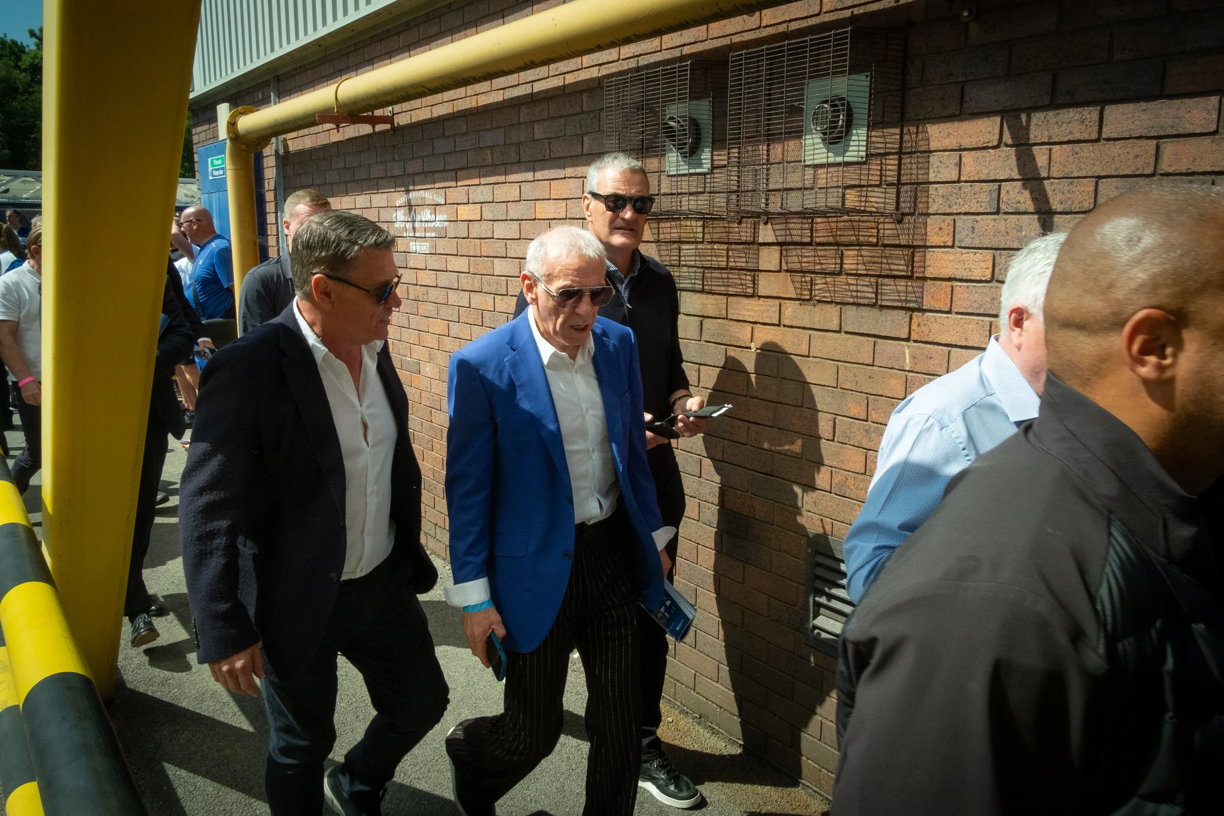 Group of men walking outside near a brick wall, with some wearing suits and sunglasses.