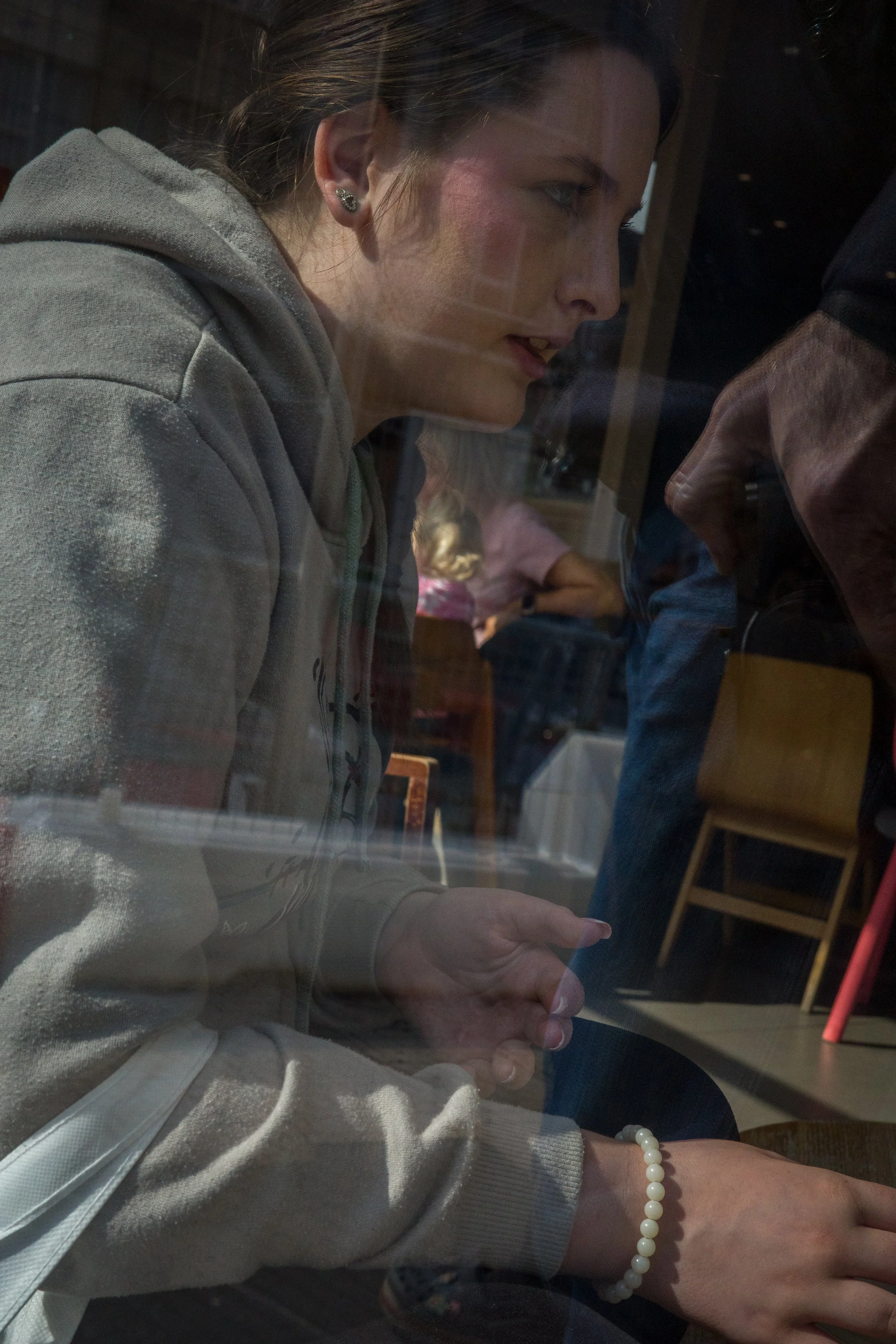 A woman with earrings and a pearl bracelet looking at a device behind a glass, with reflections of people and furniture in the glass.