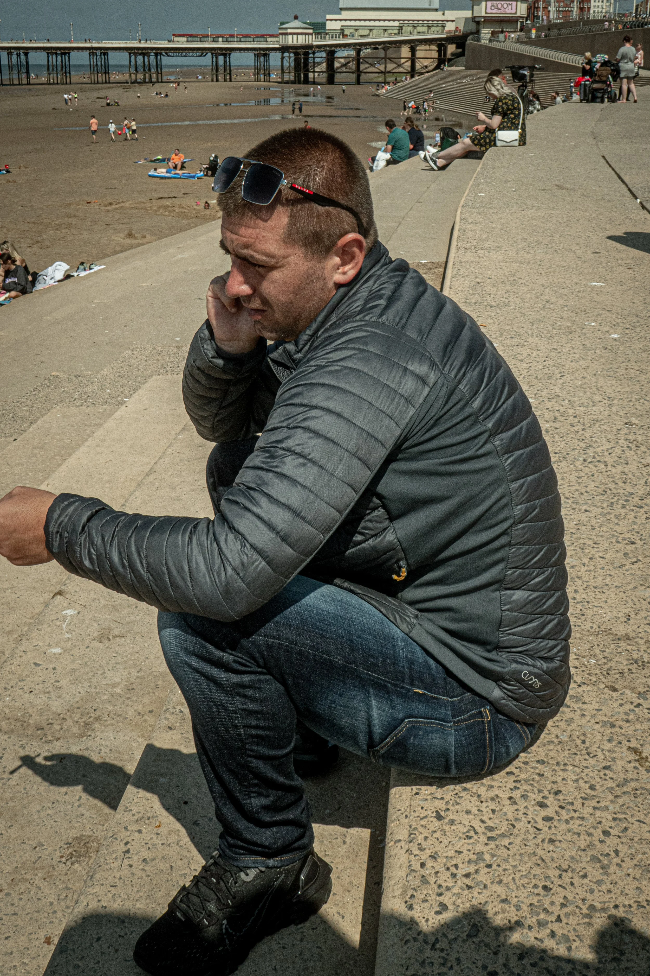 A man wearing a black jacket and jeans, sitting on a concrete bench at the beach, talking on his phone with sunglasses resting on his head. In the background, there are people on the sandy beach and a pier extending into the water, with a building la