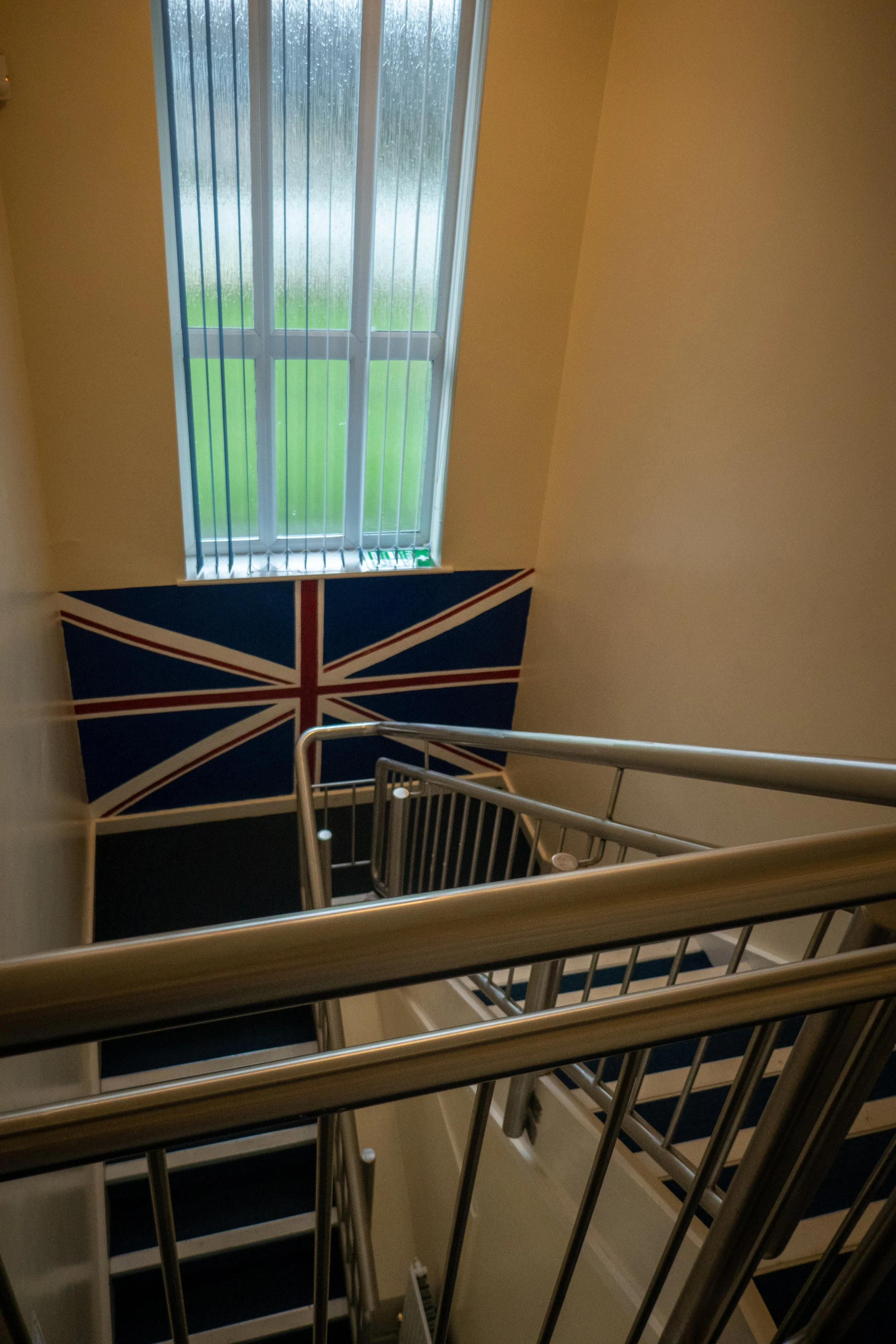 View of a stairwell with metal railings, a window with vertical blinds, and a Union Jack flag on the wall.