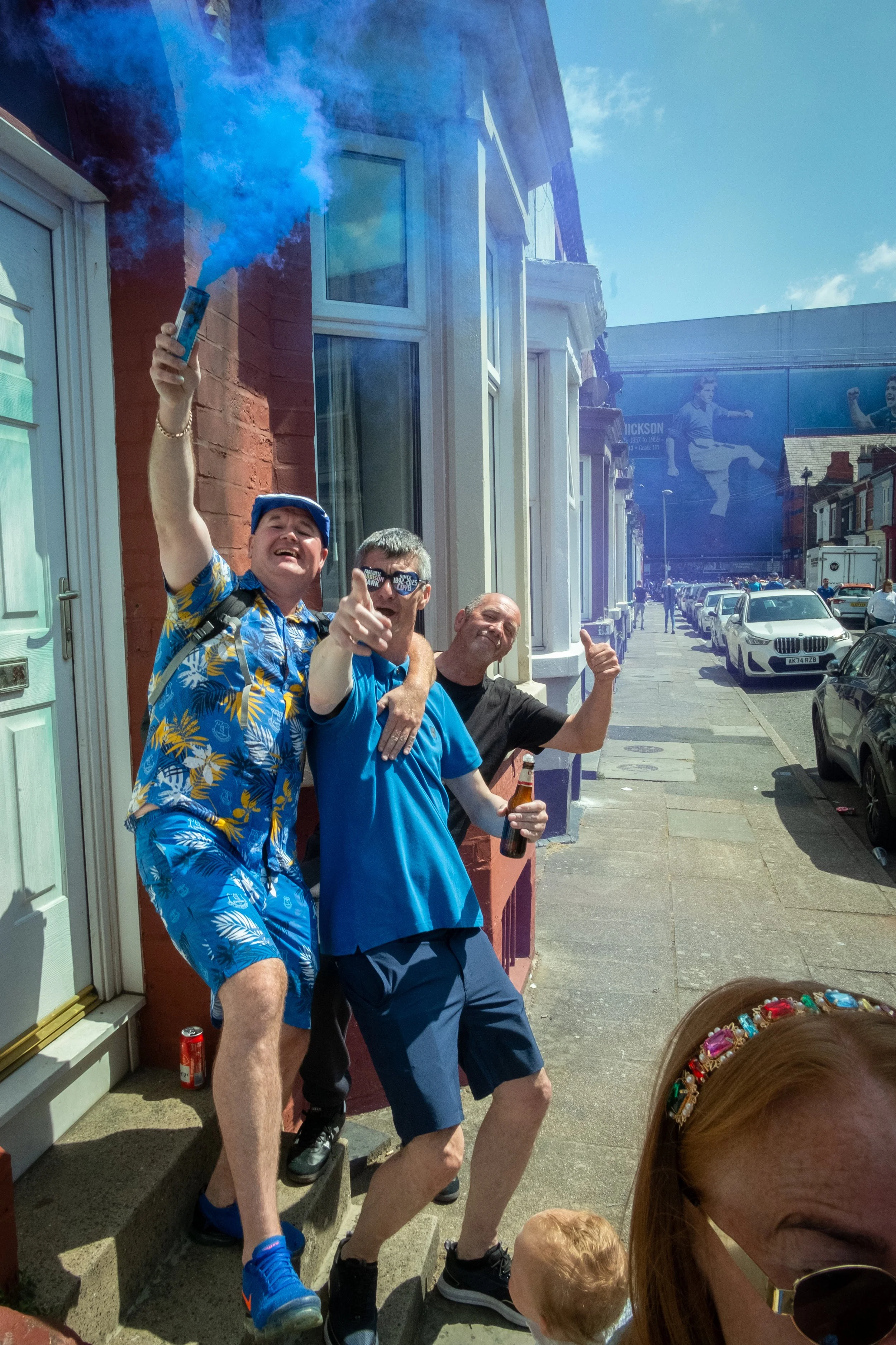 Three men celebrating outdoors, one holding a cigarette with blue smoke, one wearing sunglasses and a blue shirt, another giving a thumbs-up, on a city sidewalk with parked cars and a large billboard.