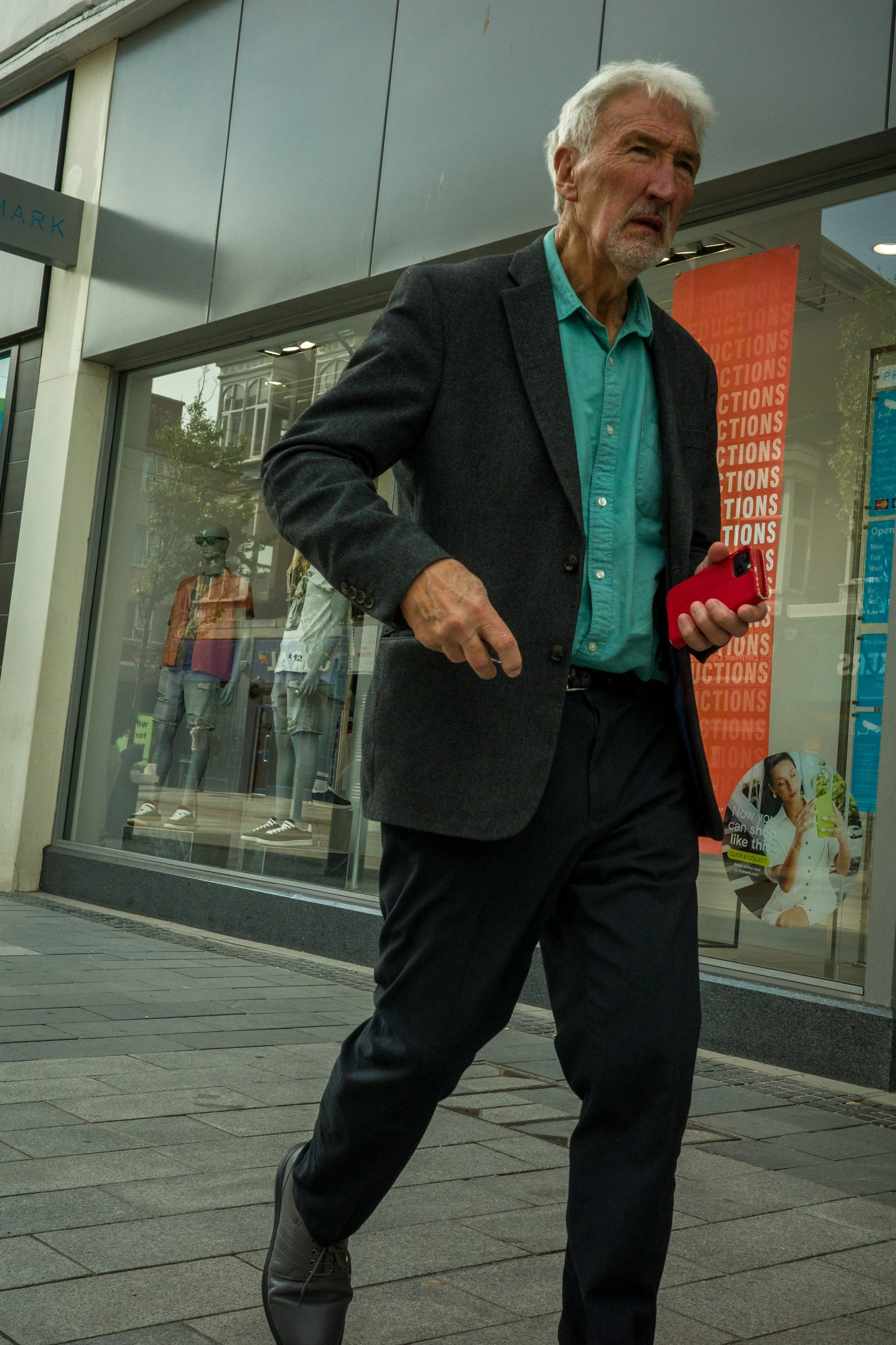 An elderly man with white hair and a beard walking on a city sidewalk, holding a red smartphone in his right hand, wearing a dark blazer, teal shirt, black pants, and gray sneakers, with store windows and mannequins in the background.