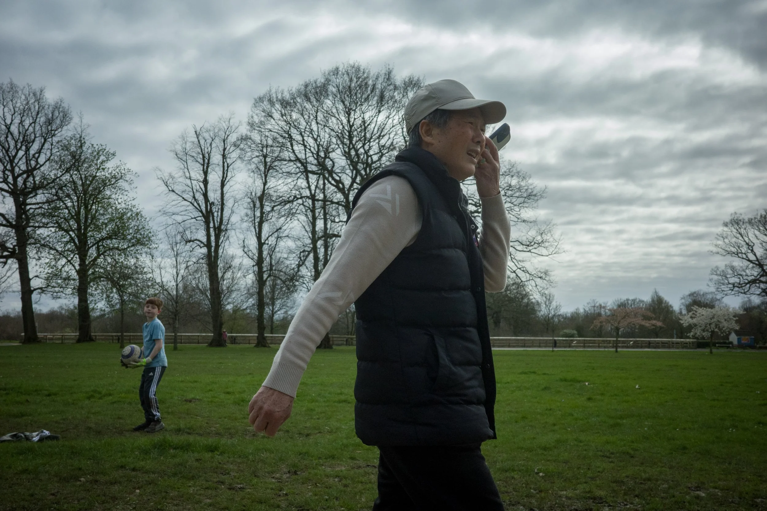 An elderly man walking outdoors on a cloudy day, wearing a beige cap, a dark vest, and a light-colored long-sleeve shirt. In the background, a young boy in a light blue shirt stands on a grass field holding a soccer ball, with leafless trees and a cl
