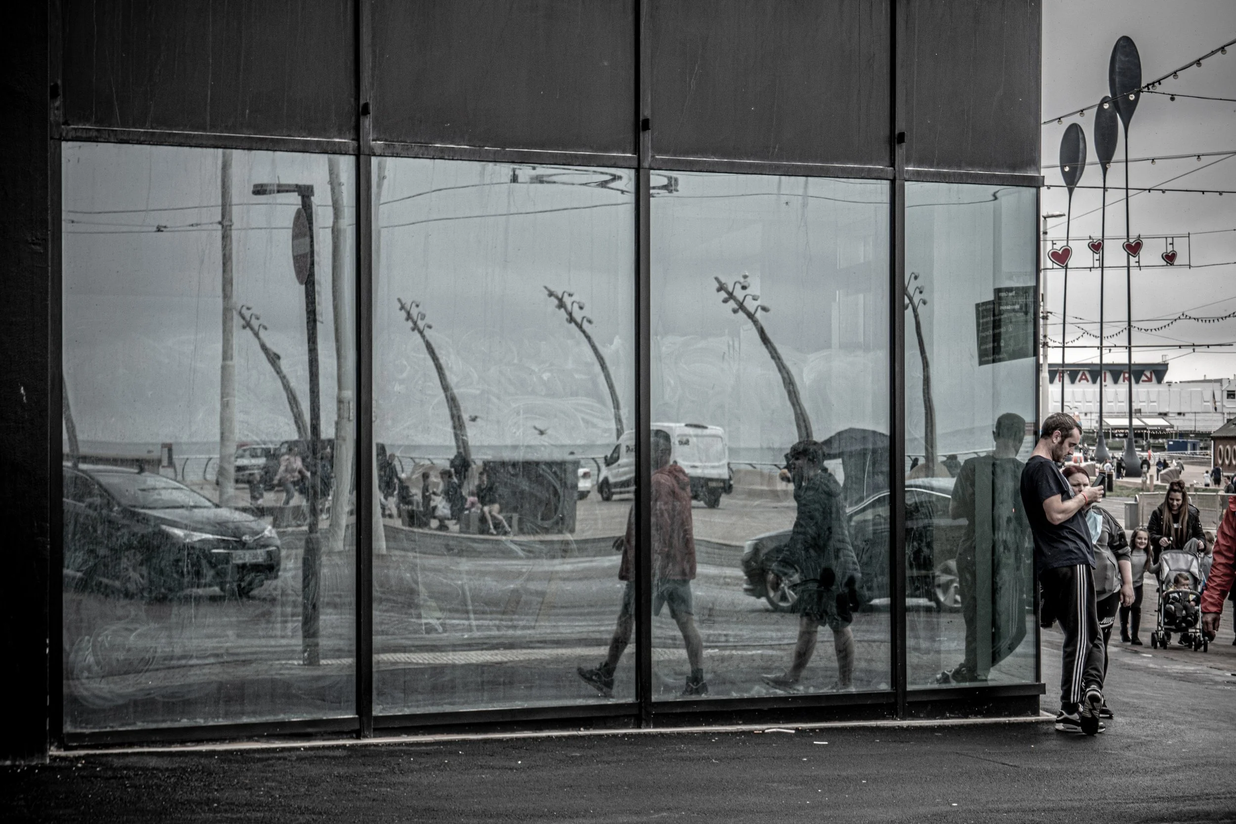 Reflected image of pedestrians walking at an outdoor area, seen through large glass windows, with some people sitting nearby and decorations hanging in the background.