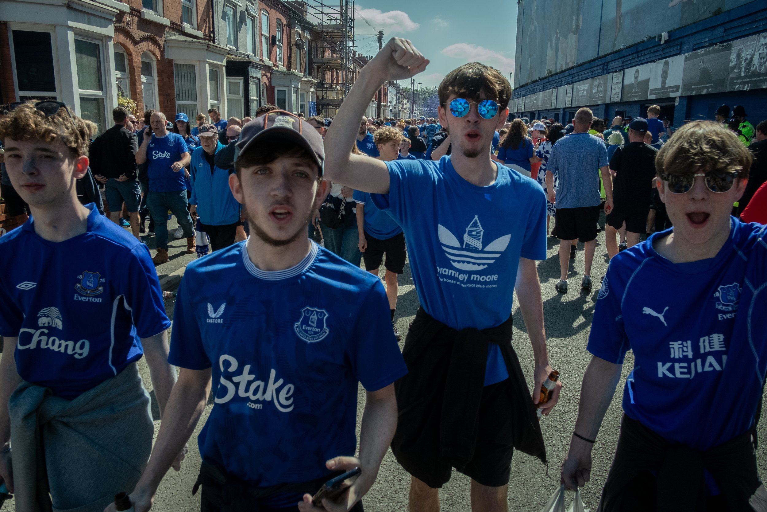 Group of young men wearing blue Everton football jerseys walking outdoors during daytime, with some raising fists and wearing sunglasses.