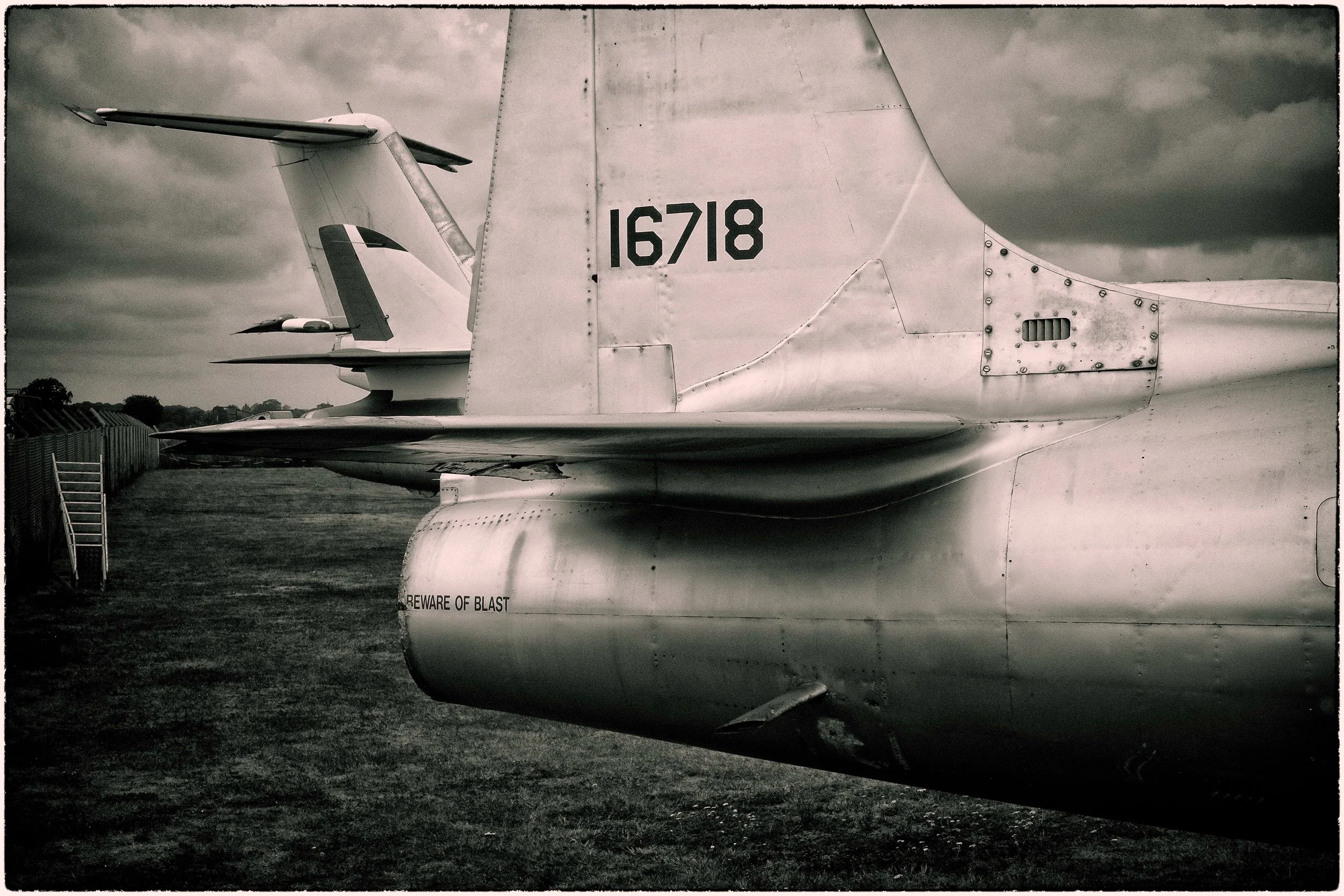 Close-up of the tail section of a military jet aircraft with the marking 16718, parked on a grassy airfield with a cloudy sky overhead.