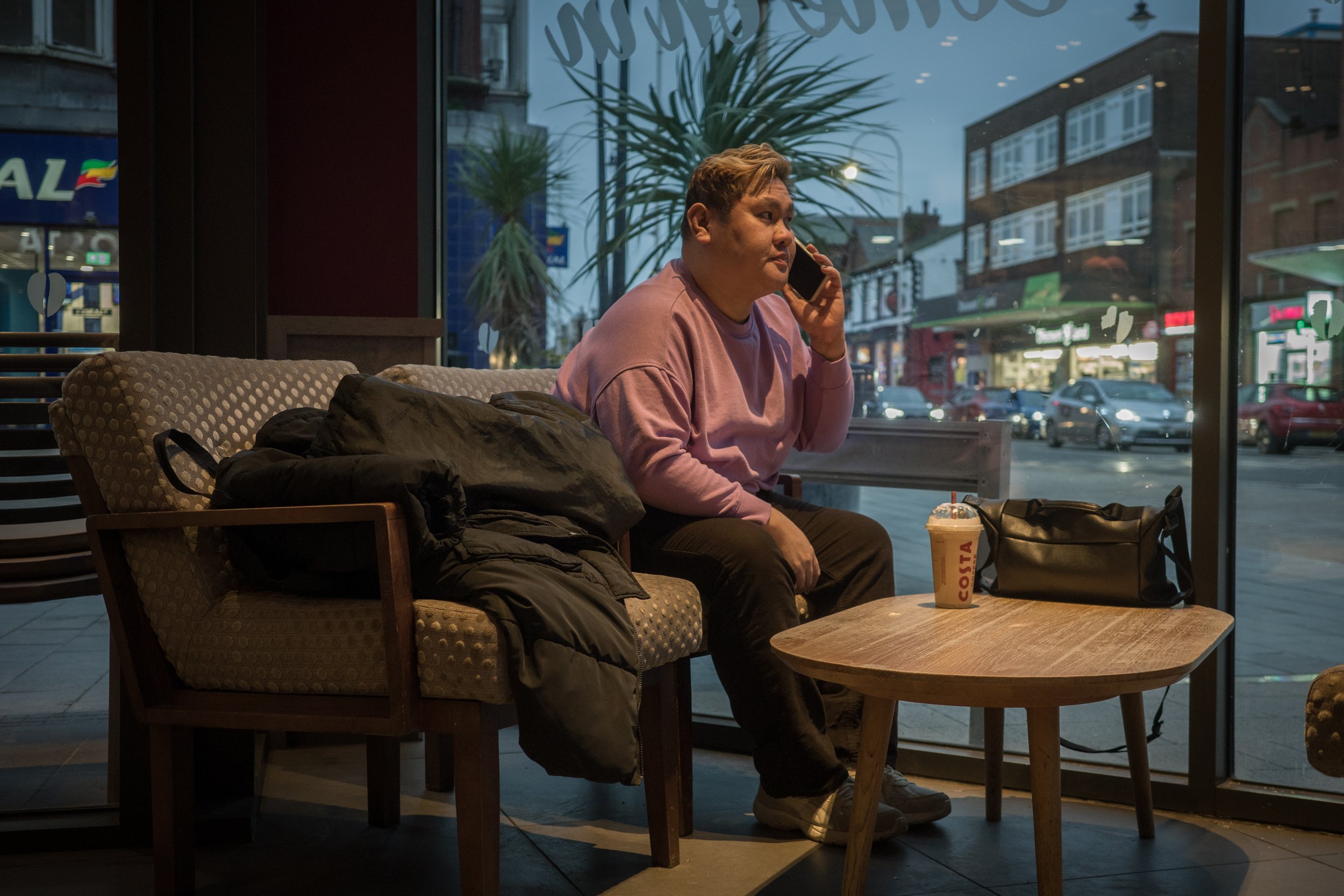 A man with light brown hair wearing a pink sweatshirt sitting inside a cafe by the window, talking on his phone. There is a Starbucks drinks cup on the table in front of him, and a black backpack on the chair beside him. Outside the window, cars are 