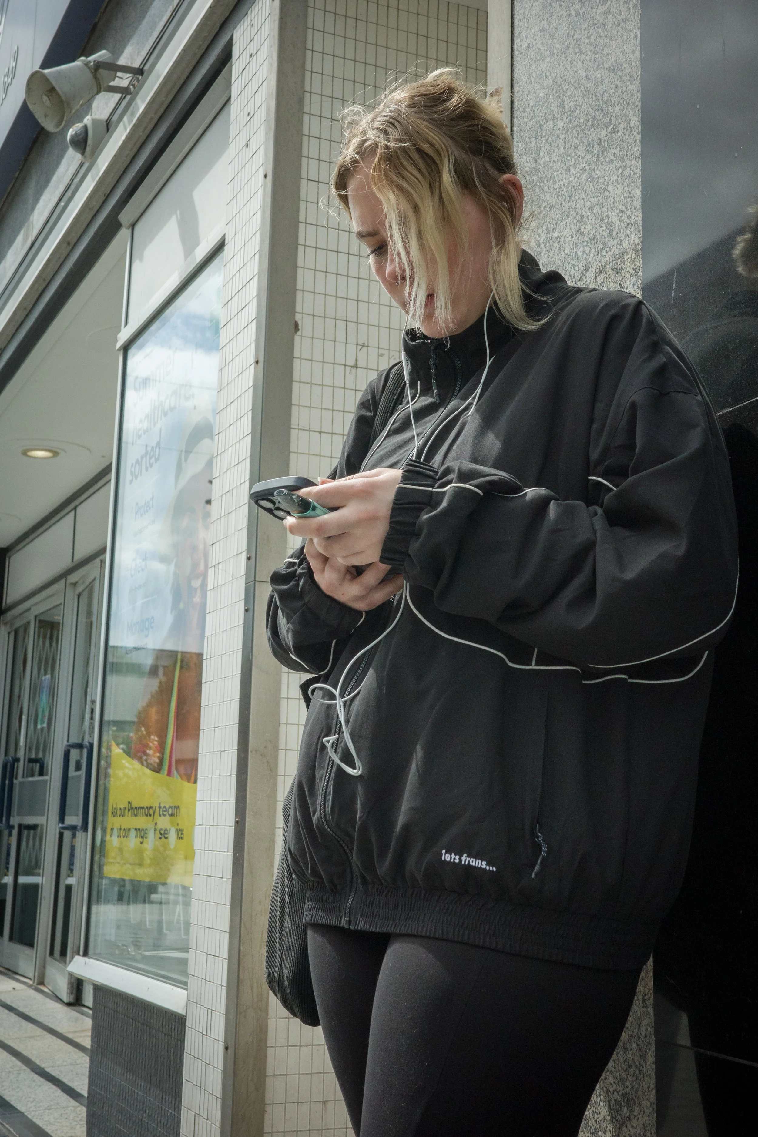 A woman with blonde hair tied back, wearing a black jacket with a zipper and black pants, is standing outside a building, looking at her phone with white earphones in her ears.