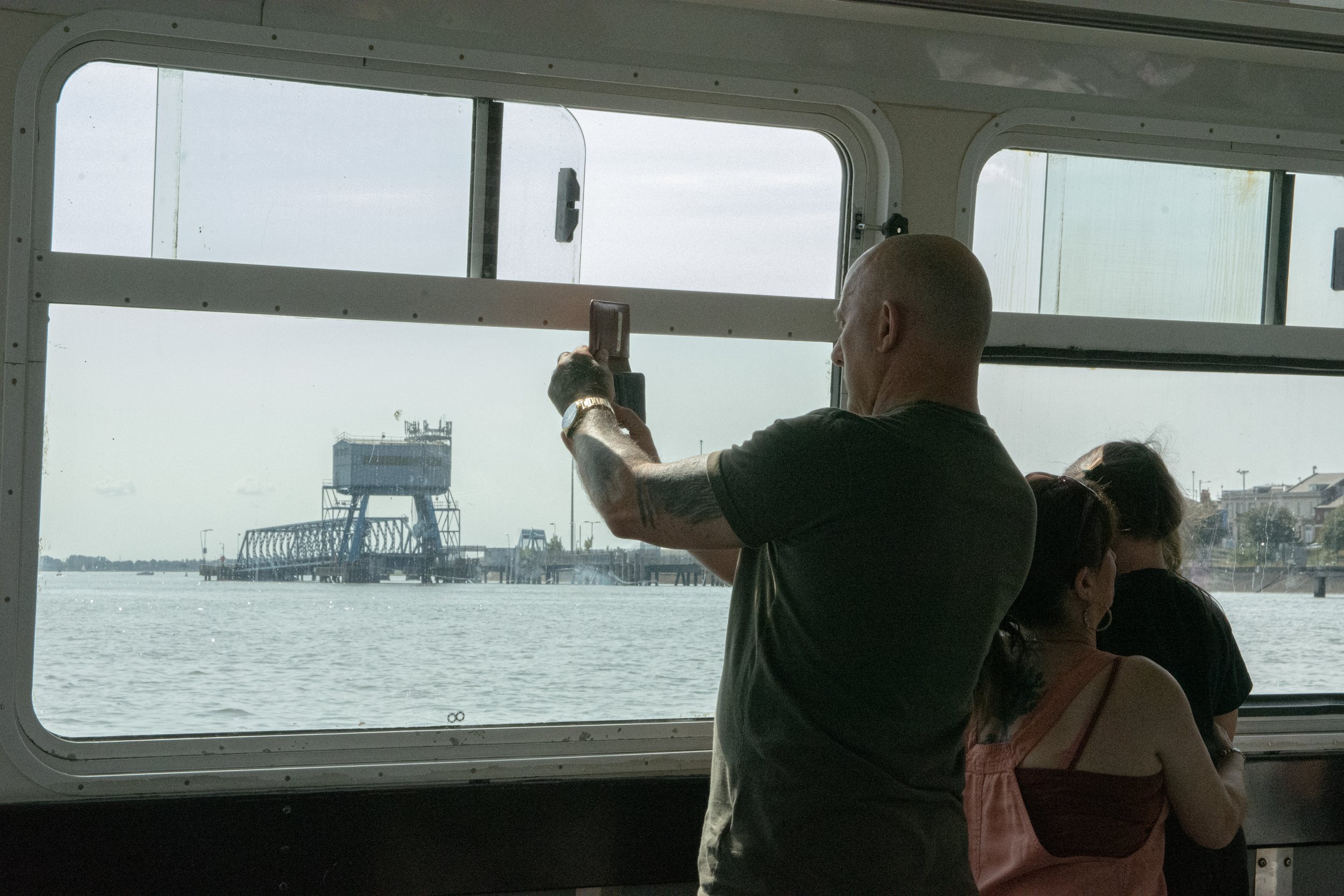 People observing the water and a bridge through a window on a ferry or boat, with an industrial structure and cityscape in the background.