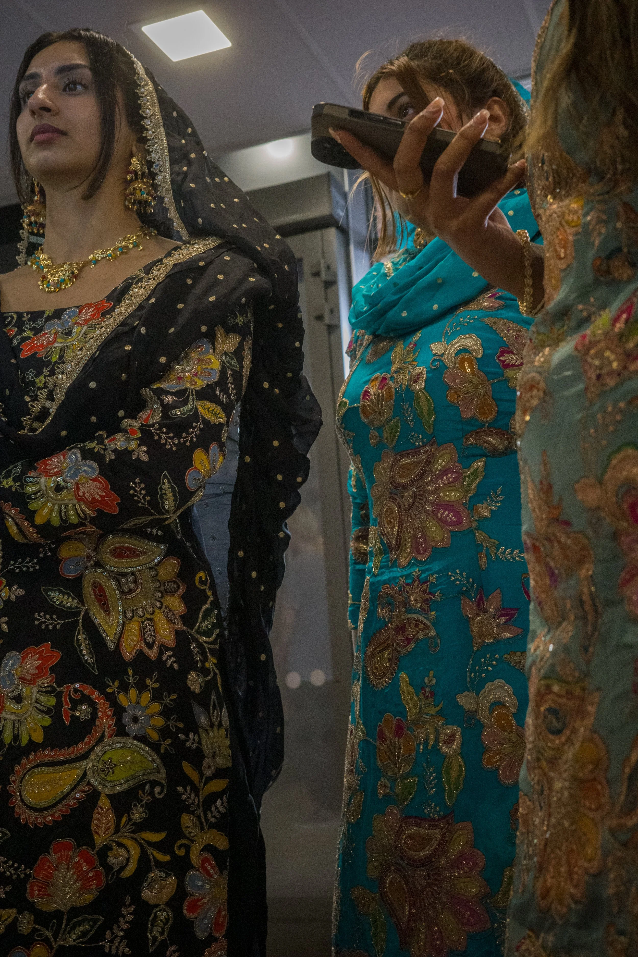 Three women wearing traditional South Asian embroidered dresses. The woman on the left is wearing a black dress with gold, red, and blue floral embroidery, and traditional jewelry. The woman in the middle, holding a cellphone, is dressed in a teal em