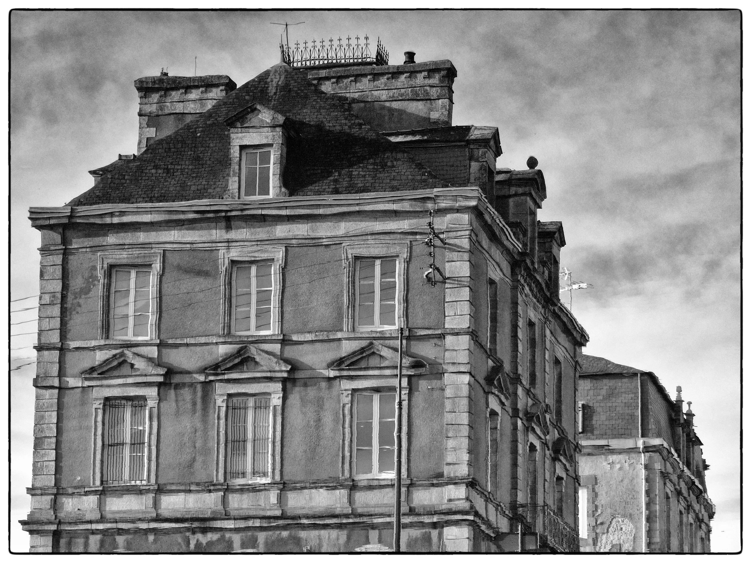 Black and white photo of an old, multi-story building with detailed stonework and multiple windows, featuring a sloped roof and decorative roof fixtures.