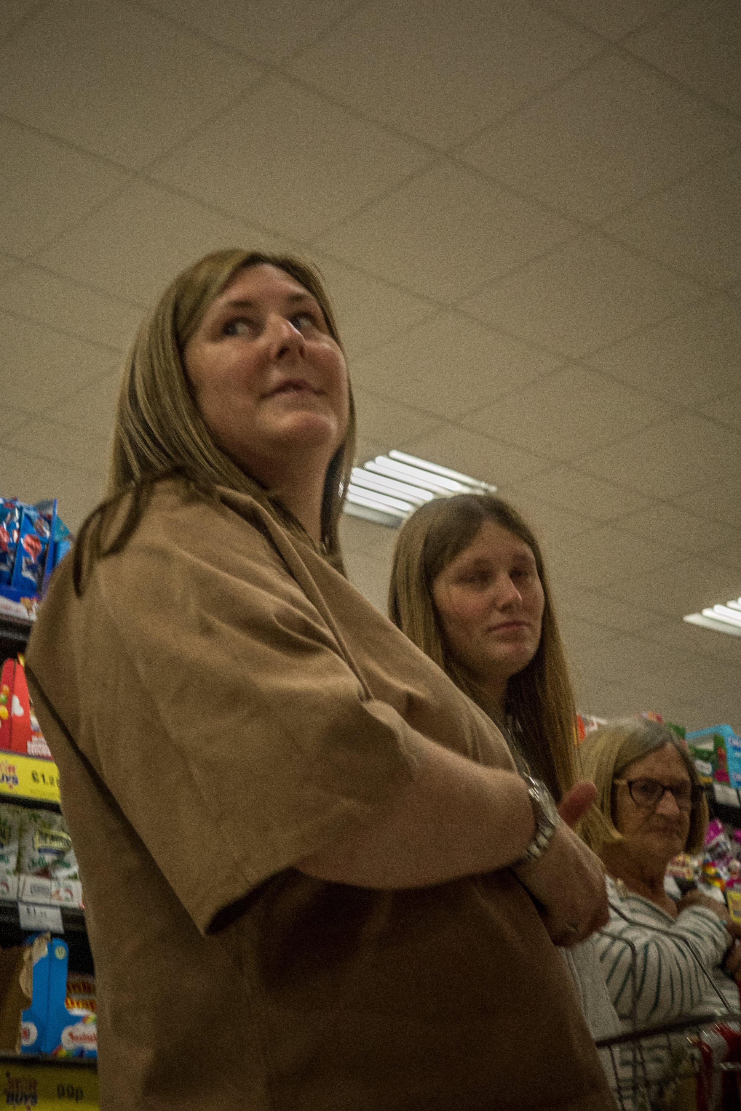 Three women standing in a grocery store aisle, one in the foreground with crossed arms, and shelves of snack products behind them.