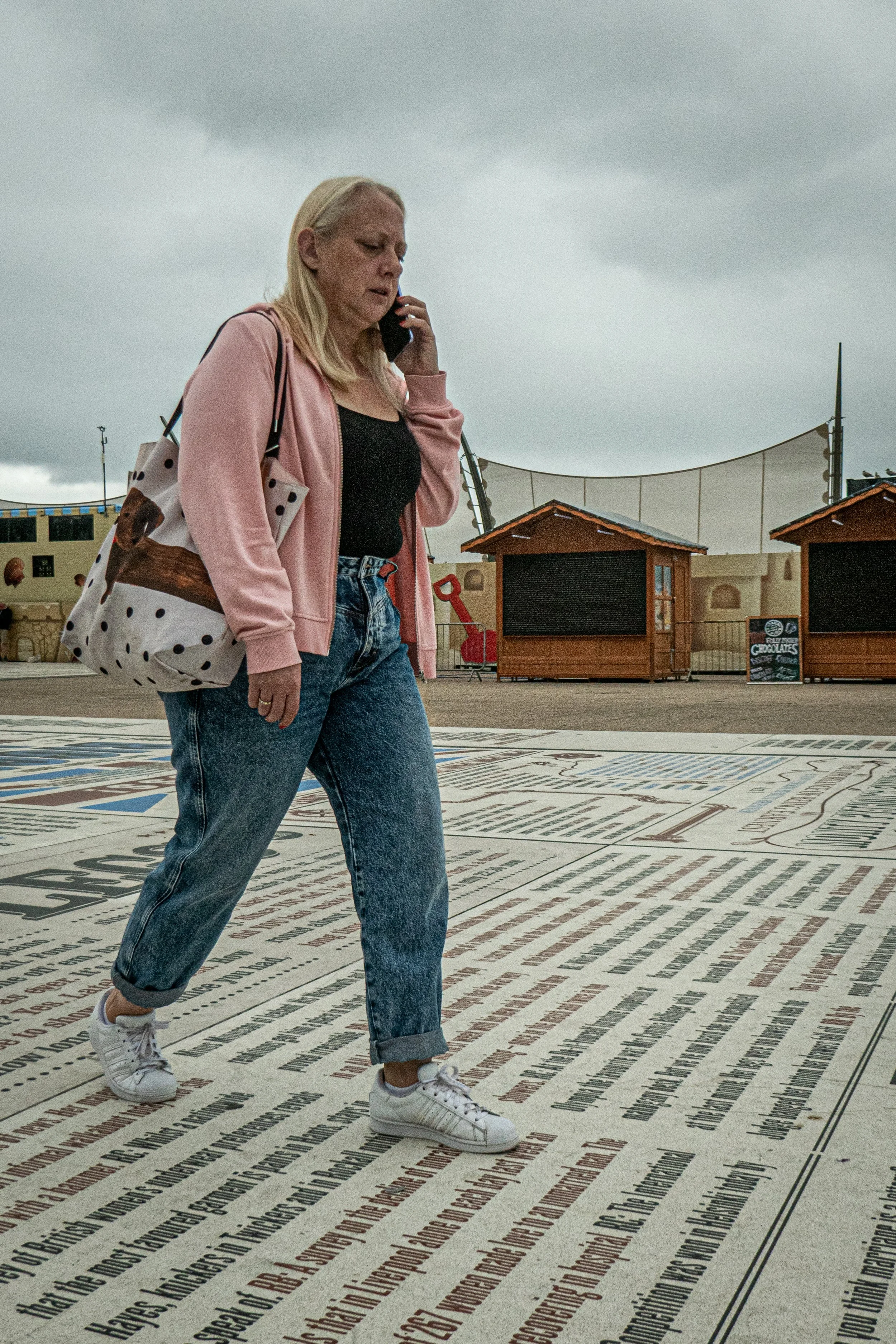 A woman in a pink hoodie and jeans walking on a surface covered with text, while talking on a cellphone, with dark cloudy skies overhead.