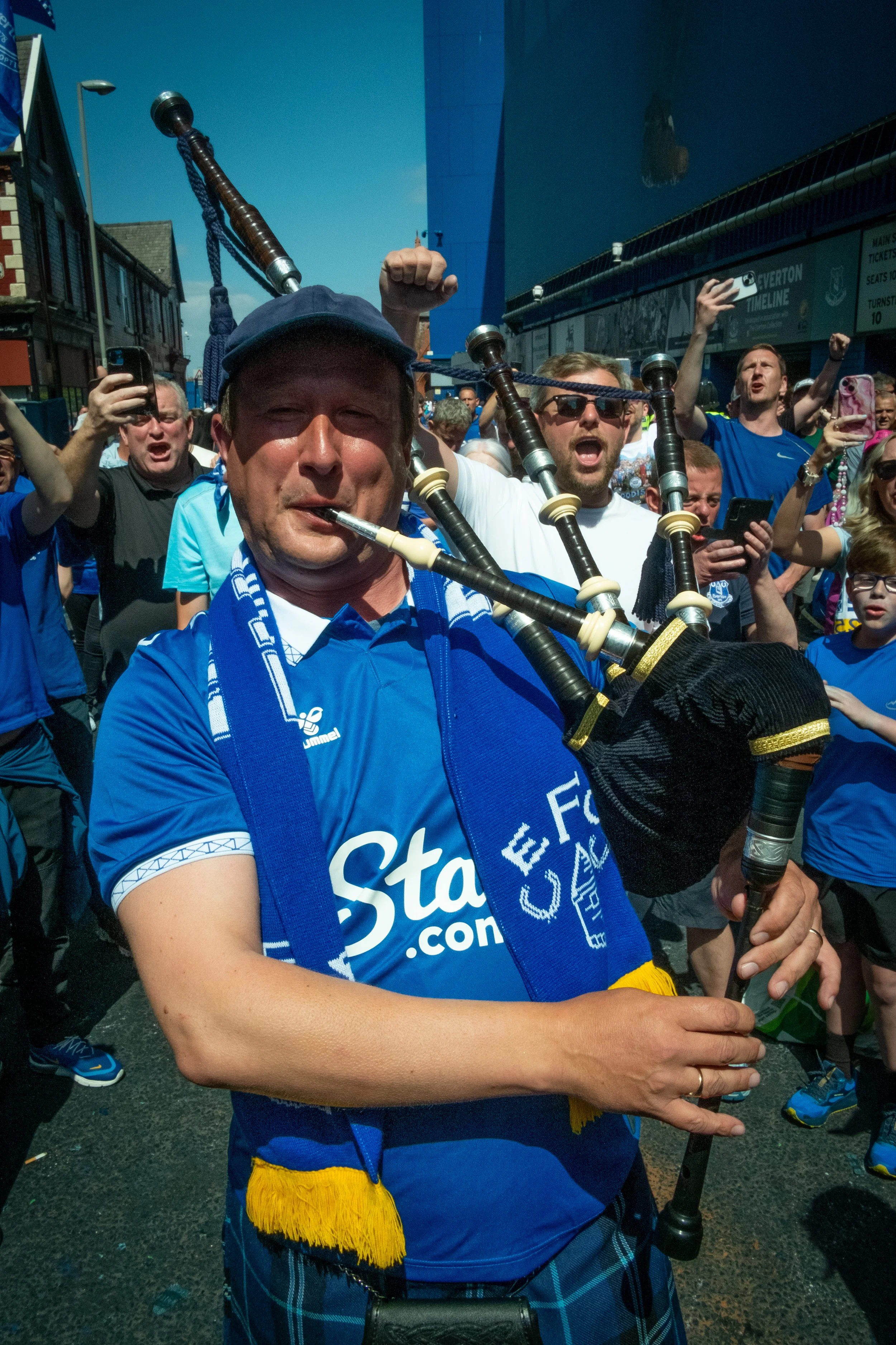 Man playing bagpipes at a crowded outdoor event, many people wearing blue shirts and cheering.